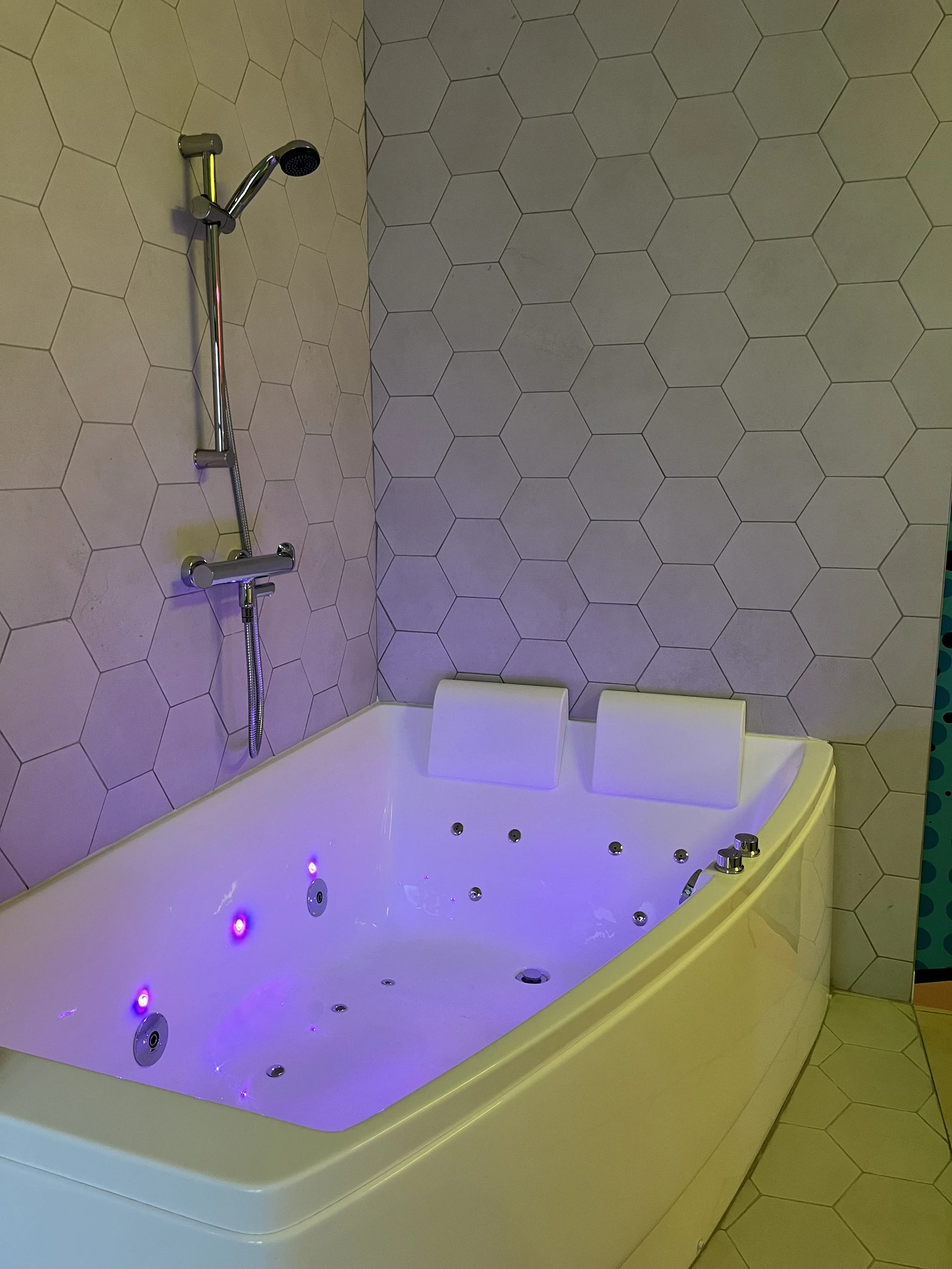 Baignoire à jets avec deux appuie-têtes, éclairée par des lumières violettes, dans une salle de bain tapissée de carreaux hexagonaux beige.
