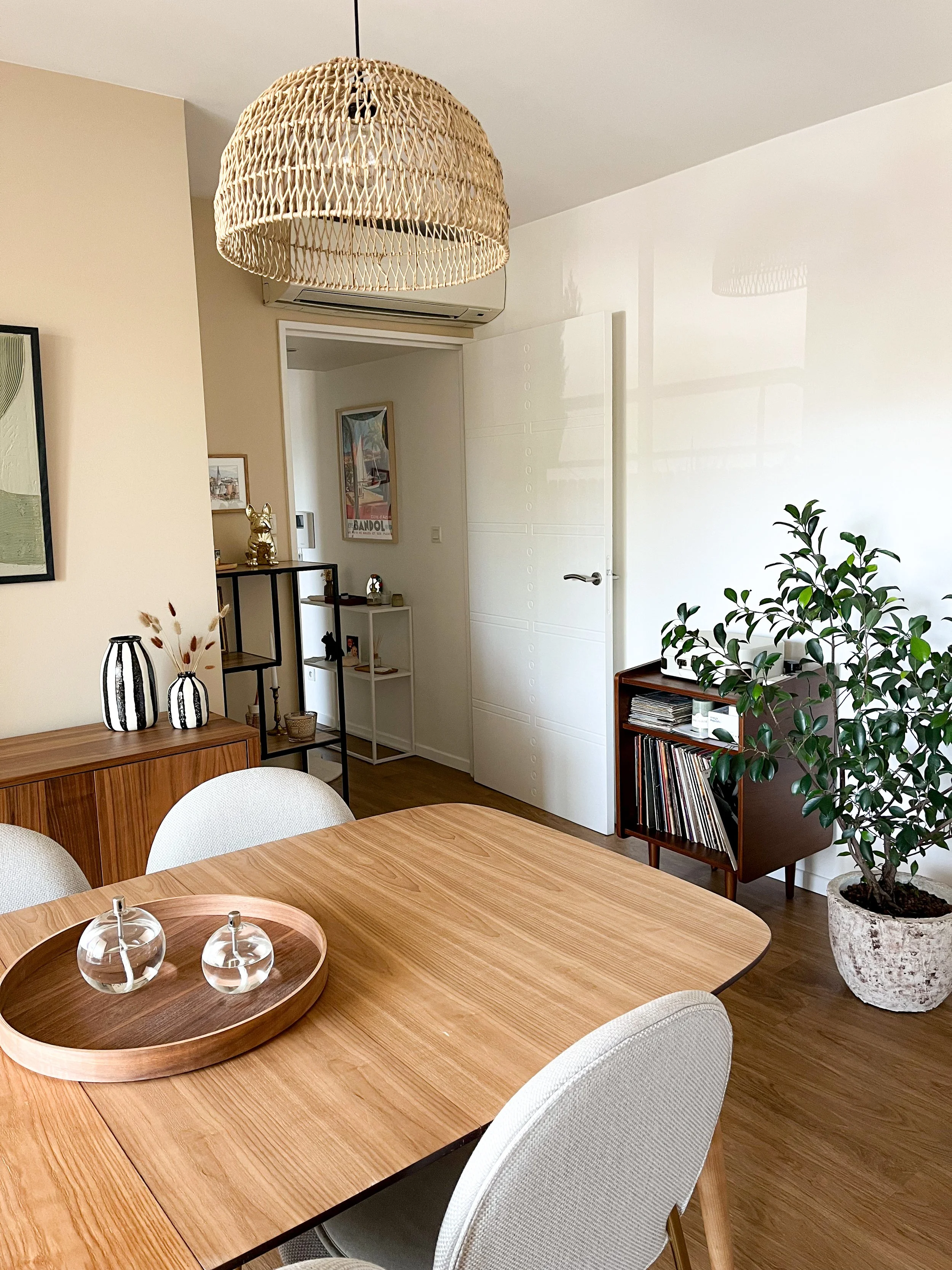 Une pièce de style contemporain avec une table en bois, des chaises beige, un grand vase en céramique avec une plante verte, une suspension en rotin au plafond, une petite étagère avec des disques vinyles, décorations et photos sur les murs.