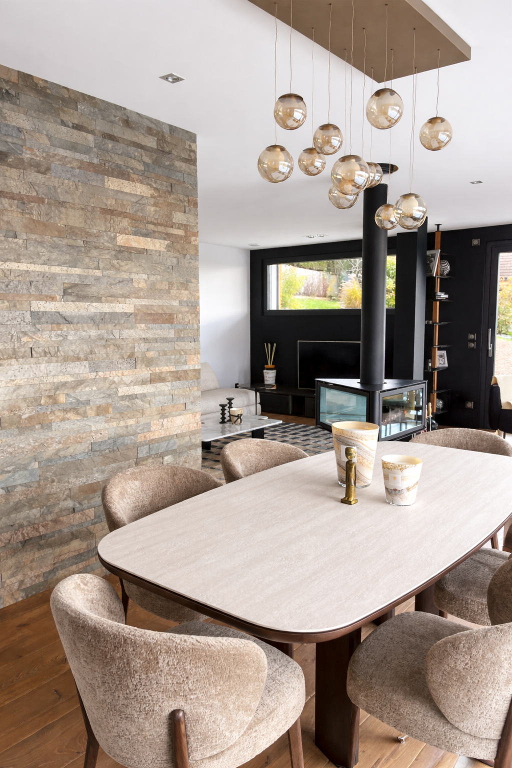 Salle à manger moderne avec table ovale et chaises en tissu beige, décoration élégante, mur en pierre apparente sur un côté, plafond avec suspension de luminaires en verre, salon avec cheminée en verre, télévision, canapé beige et pièce de décoration.