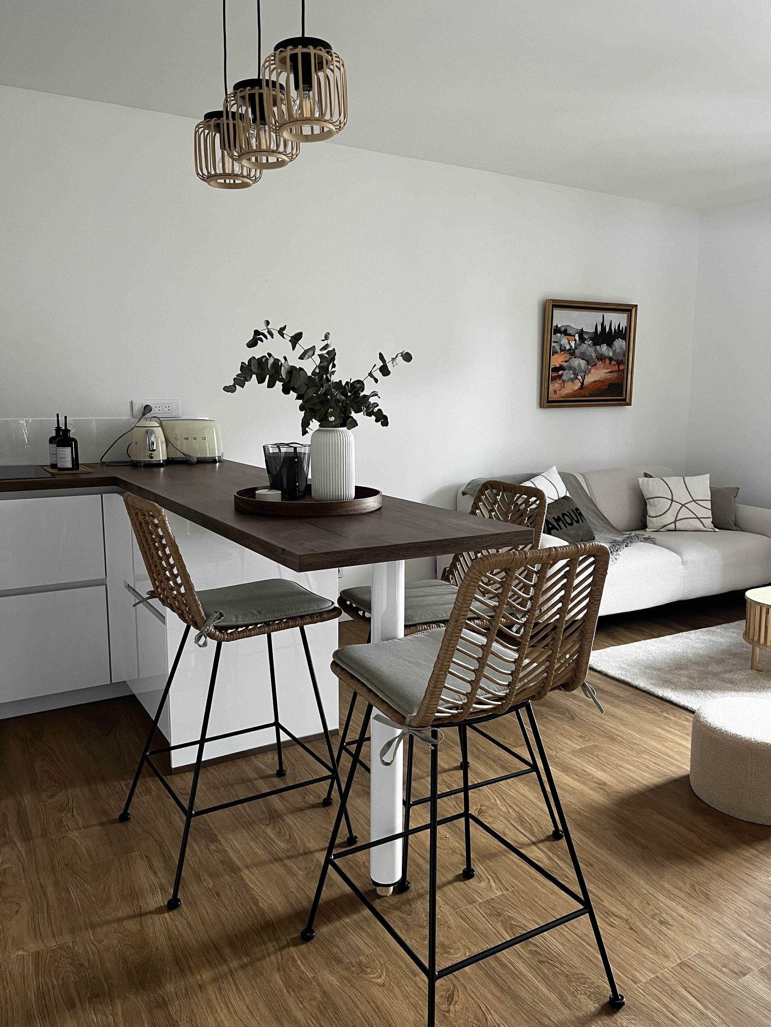 Intérieur moderne d'un salon avec table de bar en bois, chaises en rotin avec coussins, canapé blanc, tableau sur le mur, lampes suspendues en bois, tapis beige, et éléments de cuisine en arrière-plan.