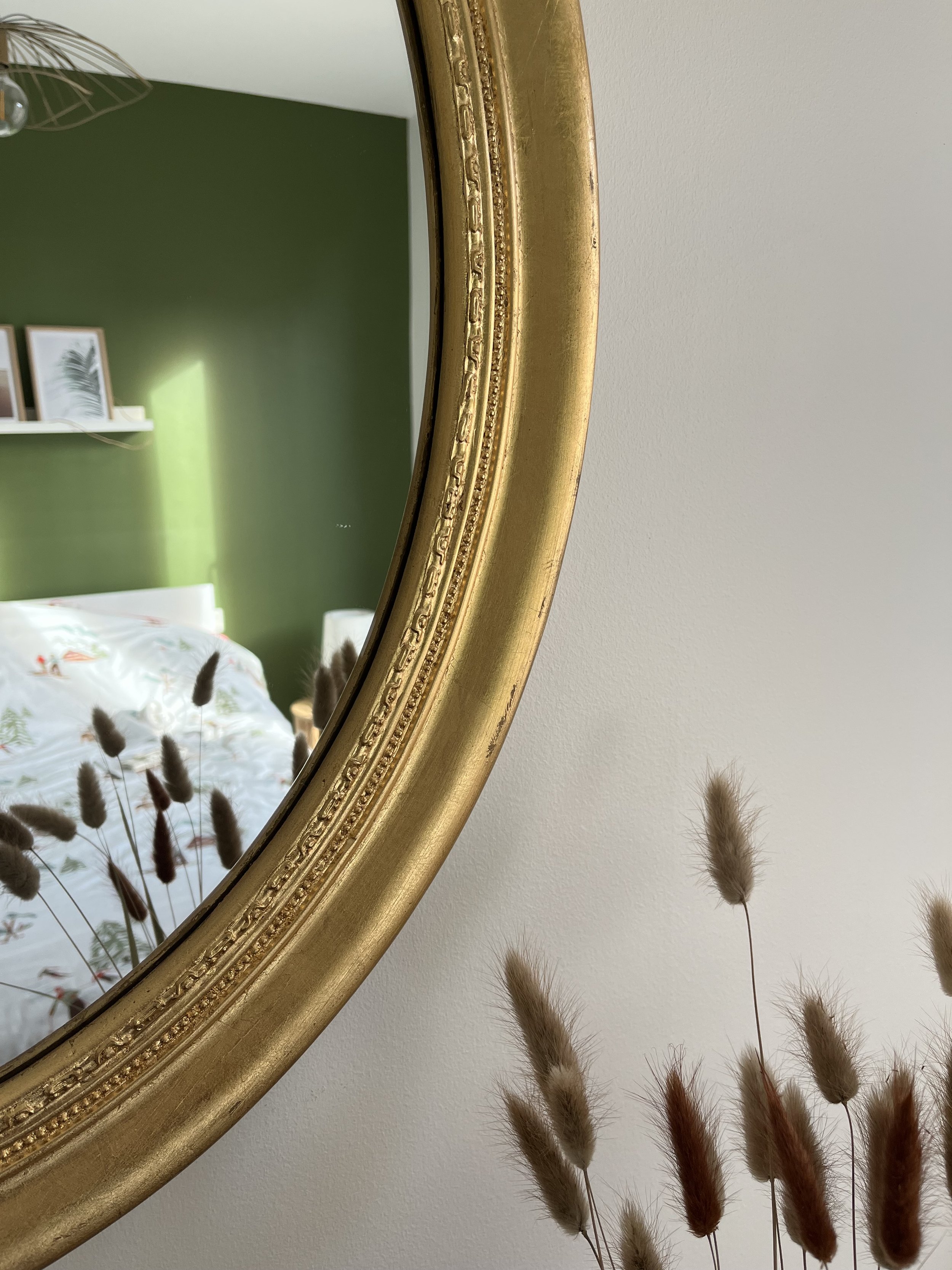 Un miroir doré rond accroché à un mur blanc, reflétant une chambre avec un mur vert, une étagère blanche avec des cadres, et un lit avec une couette à motifs, avec des plantes séchées devant le miroir.