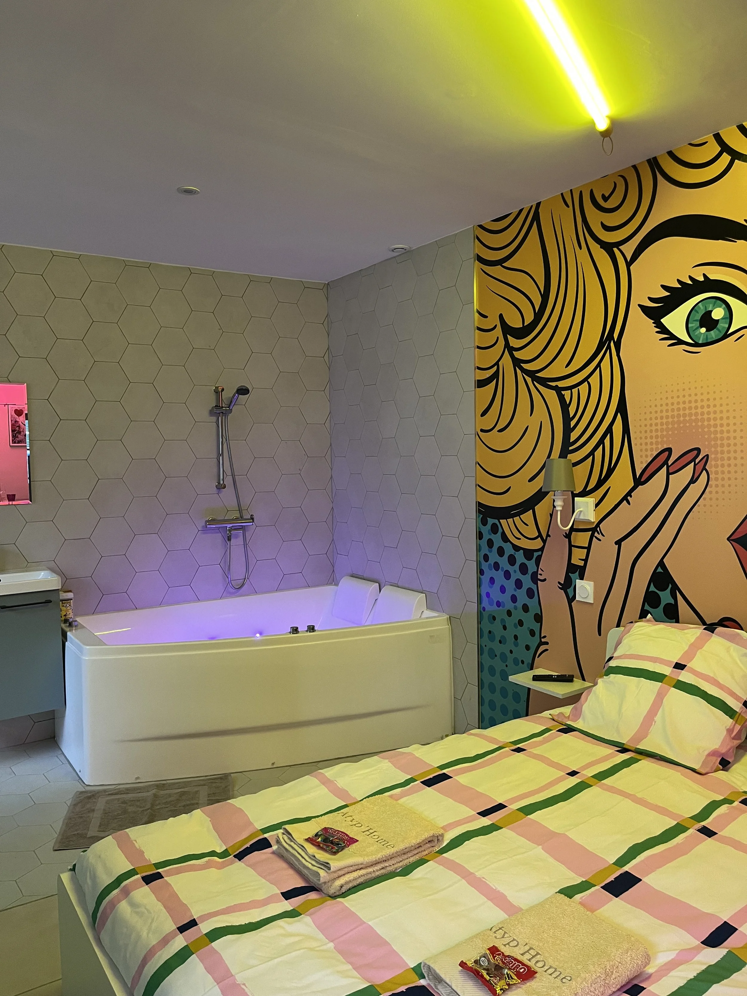 Chambre avec lit à carreaux roses, verts, jaunes, et blancs. Mur en mosaïque hexagonale grise. Bureau avec serviettes de bain et chocolat. Mur de style pop art représentant une femme avec cheveux blonds, yeux verts, et main près du visage. Baignoire avec jets lumineux, douche et robinet. Éclairage jaune plafond.