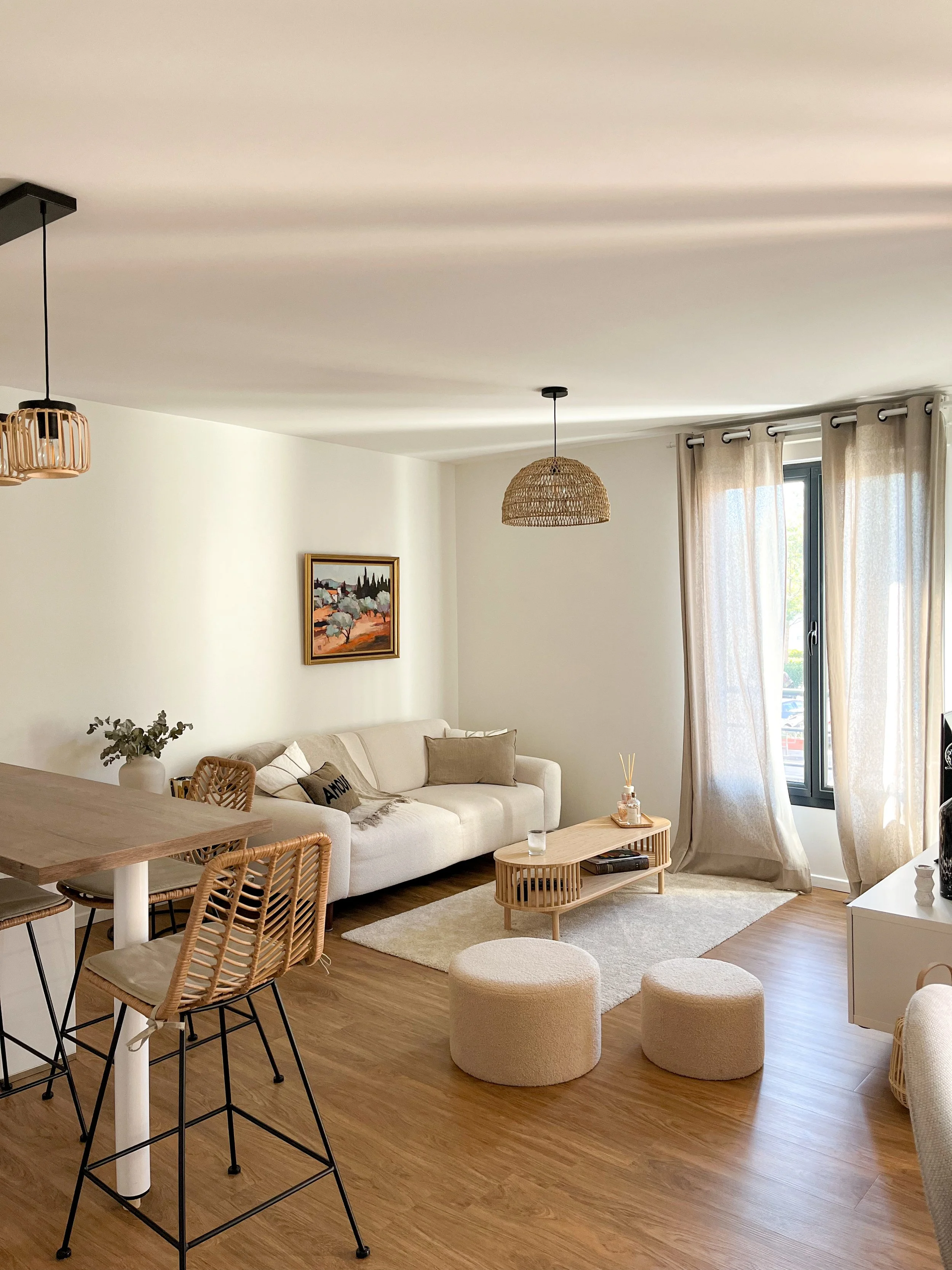 Salon moderne avec canapé beige, rideaux clairs, table basse en bois, deux poufs, lampe en osier et décoration minimaliste.