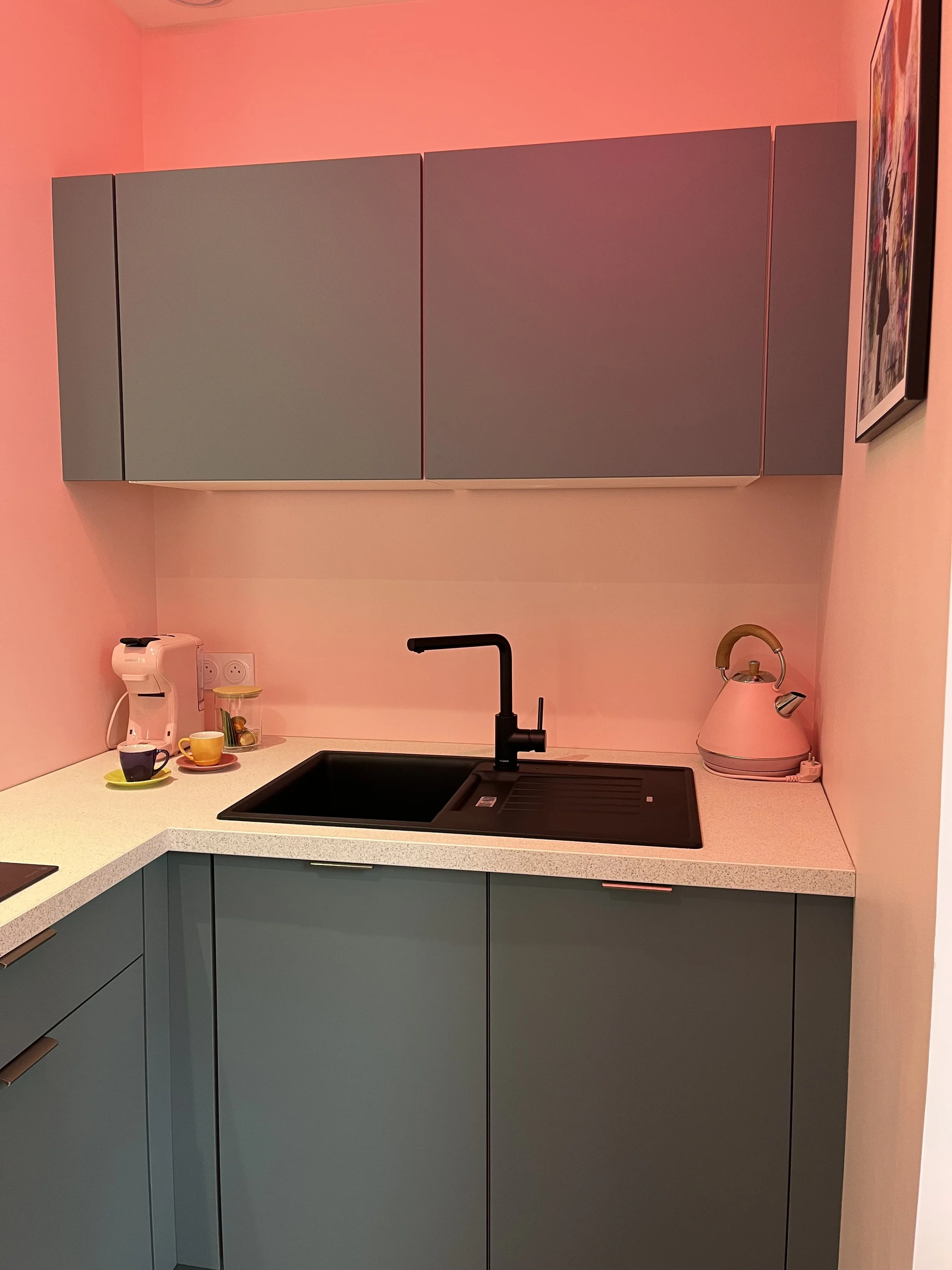 Une petite cuisine avec des meubles gris et un plan de travail en blanc, un évier noir avec robinet noir, une bouilloire rose pâle et des tasses avec soucoupes sur le comptoir, une machine à café, et un tableau coloré accroché au mur.