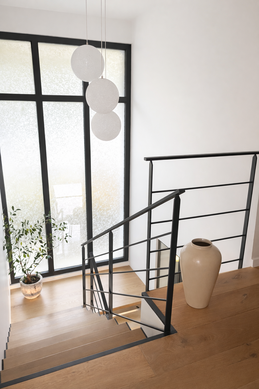 Escalier en bois avec rampe en métal noir, grande fenêtre frosted, plante en pot, grande vase beige sur une table en bois.