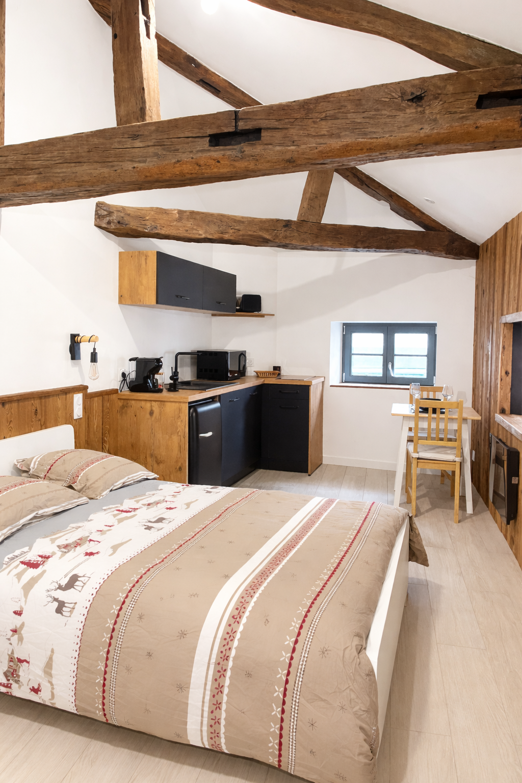 Chambre avec un lit avec une décoration de Noël, une petite cuisine en bois, une table avec deux chaises, bois apparent au plafond et sur un mur, petite fenêtre.