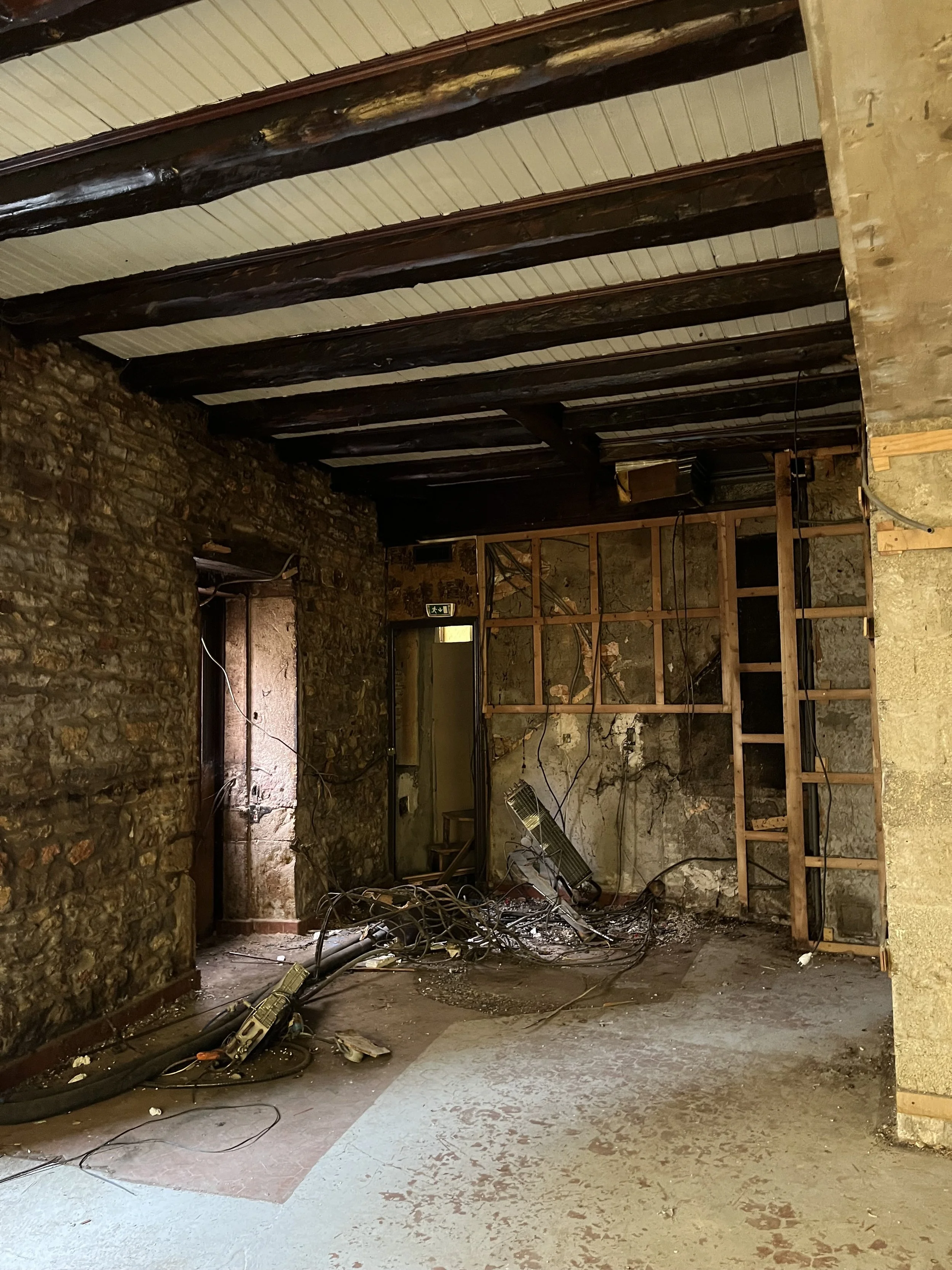 Intérieur d'une pièce en cours de rénovation, avec murs en pierre, plafond avec poutres en bois et câbles électriques dénudés au sol.