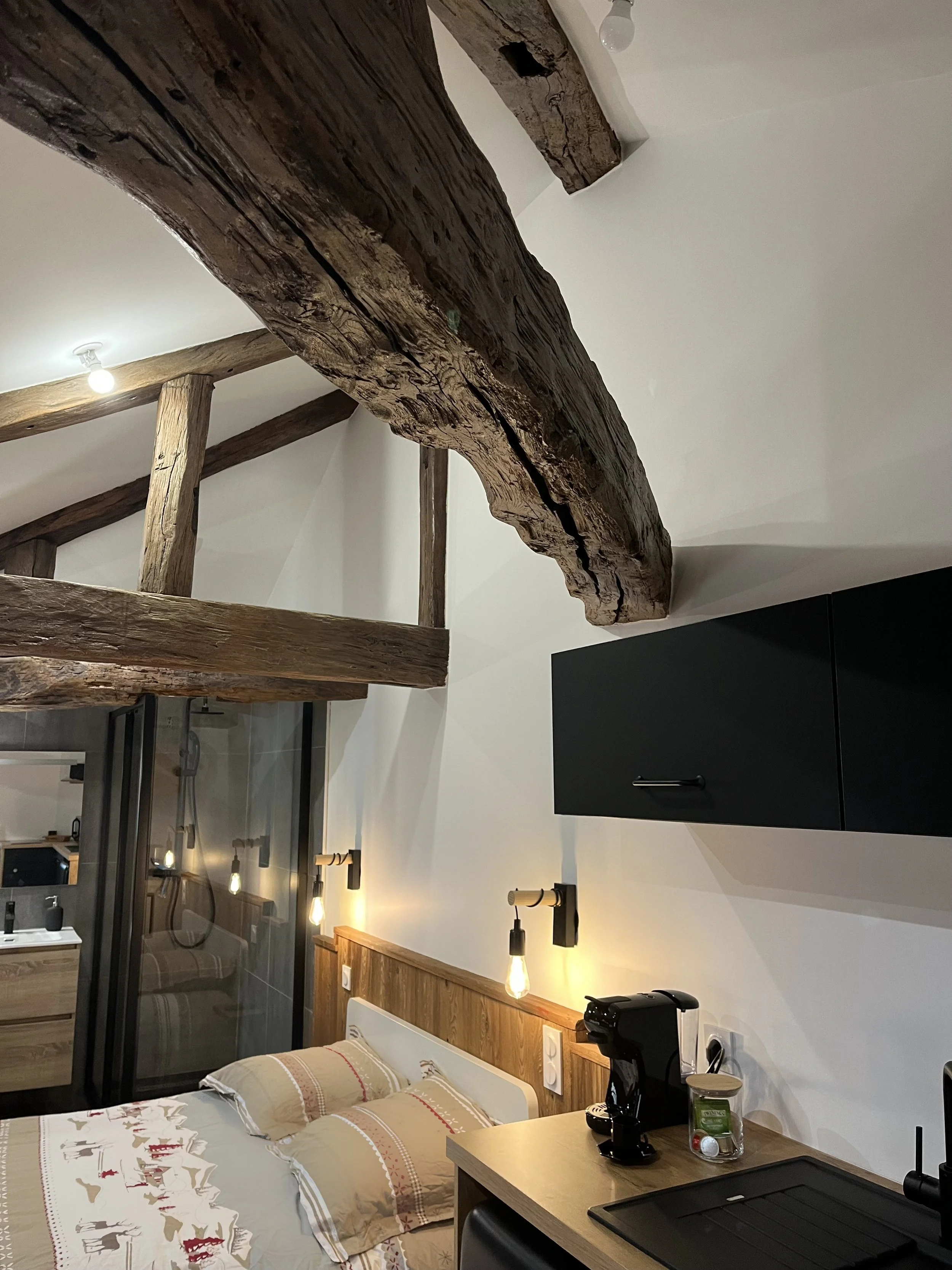 Intérieur d'une chambre avec des poutres en bois apparentes au plafond, un lit avec des oreillers décorés, une table avec une machine à café, une lampe, et un mur avec un placard noir.