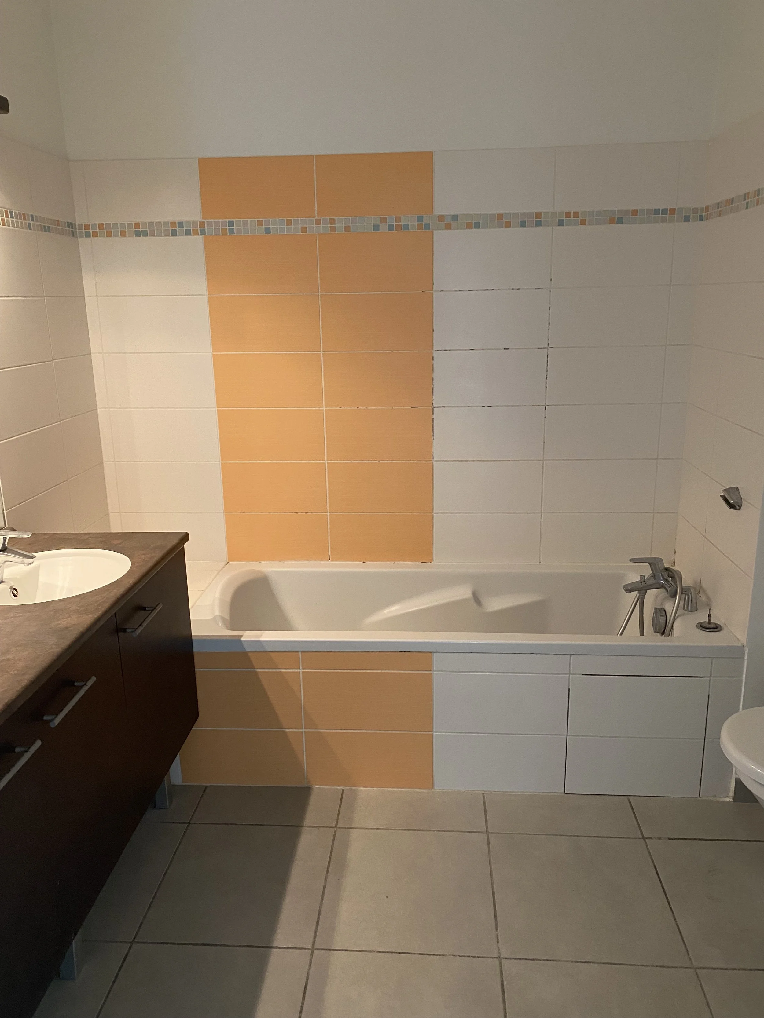 Salle de bain avec une baignoire blanche, un lavabo avec une armoire en bois foncé, des carreaux beige et orange sur le mur, et un sol en carrelage gris.