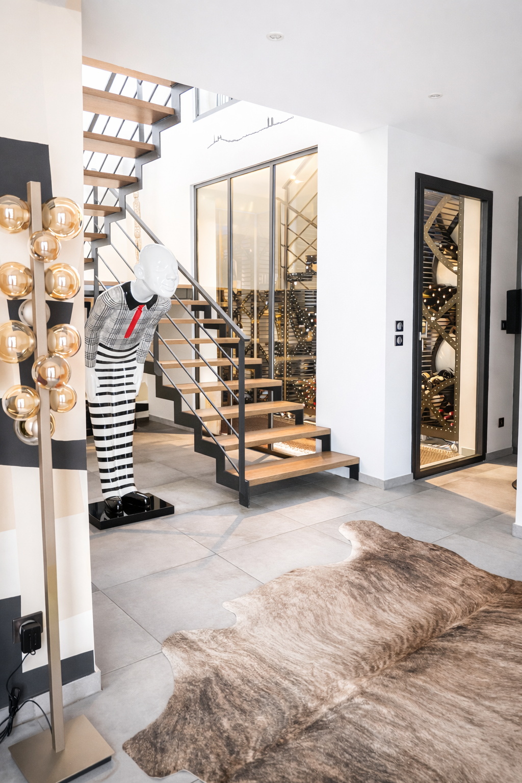Intérieur moderne d'une maison avec sculpture d'un homme en noir et blanc, escalier en bois et métal, étagère à vin, tapis en peau de vache et décorations festives, probablement pour Noël.