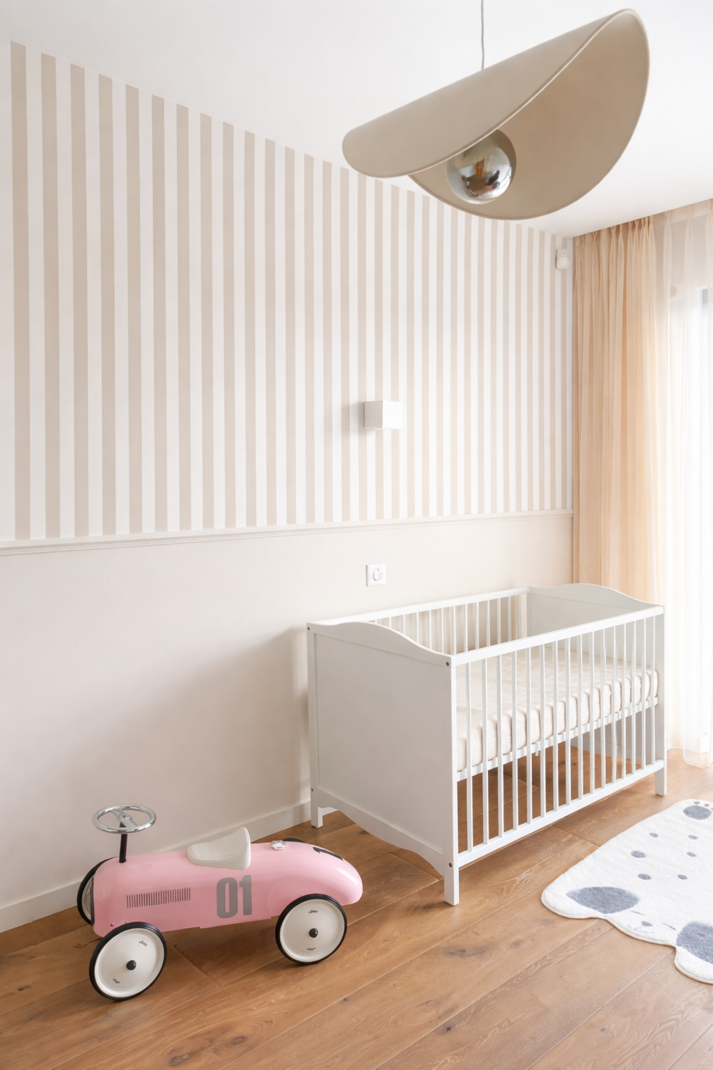 Chambre de bébé avec berceau blanc, jouet voiture rose, tapis de sol blanc avec motifs de cailloux, mur à rayures beige et blanc, rideaux beige clair, suspension moderne
