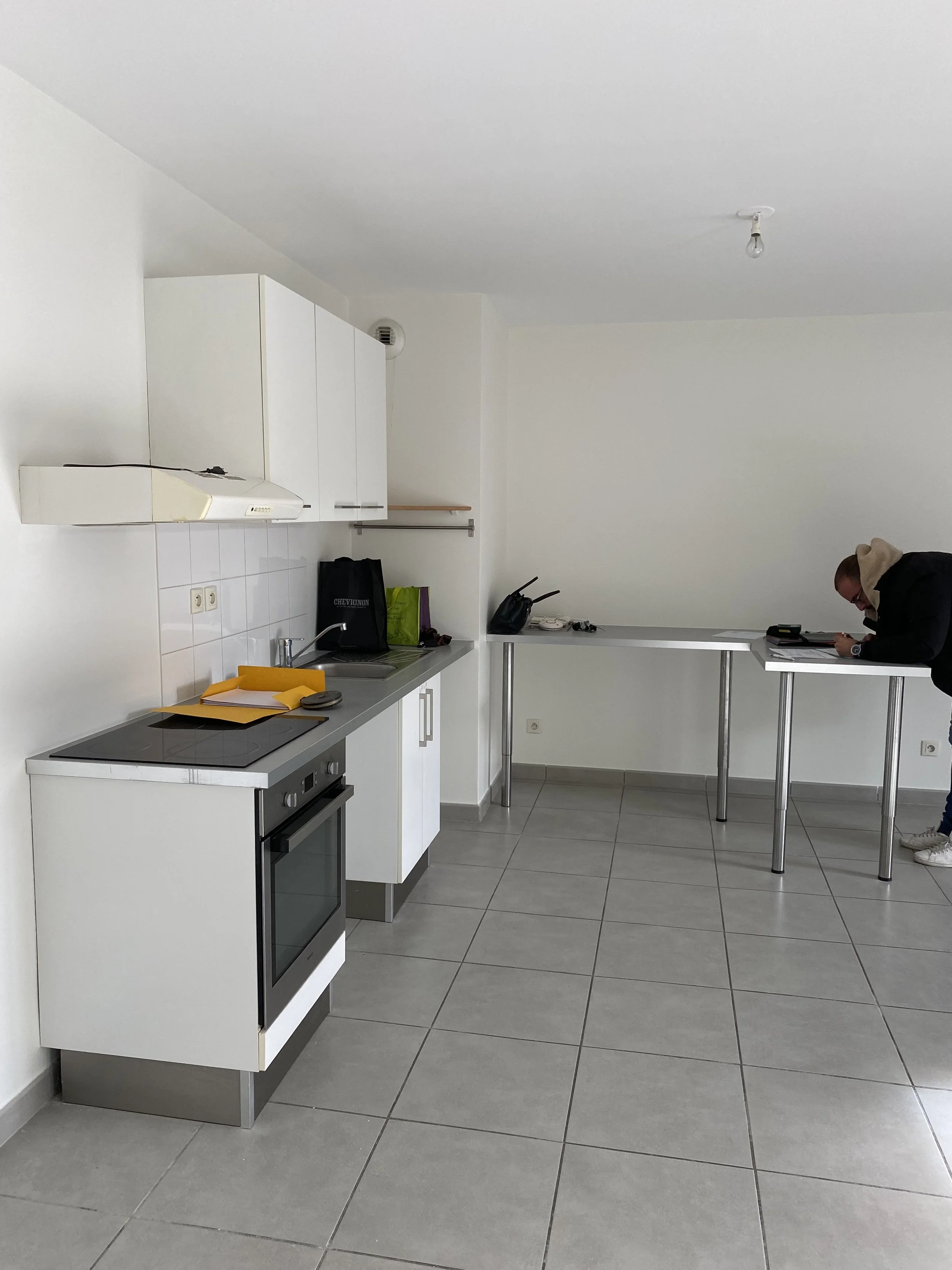 Cuisine moderne avec meubles blancs, cuisinière électrique, plans de travail avec des dossiers, un sac et une personne penchée sur une table avec des objets dessus.