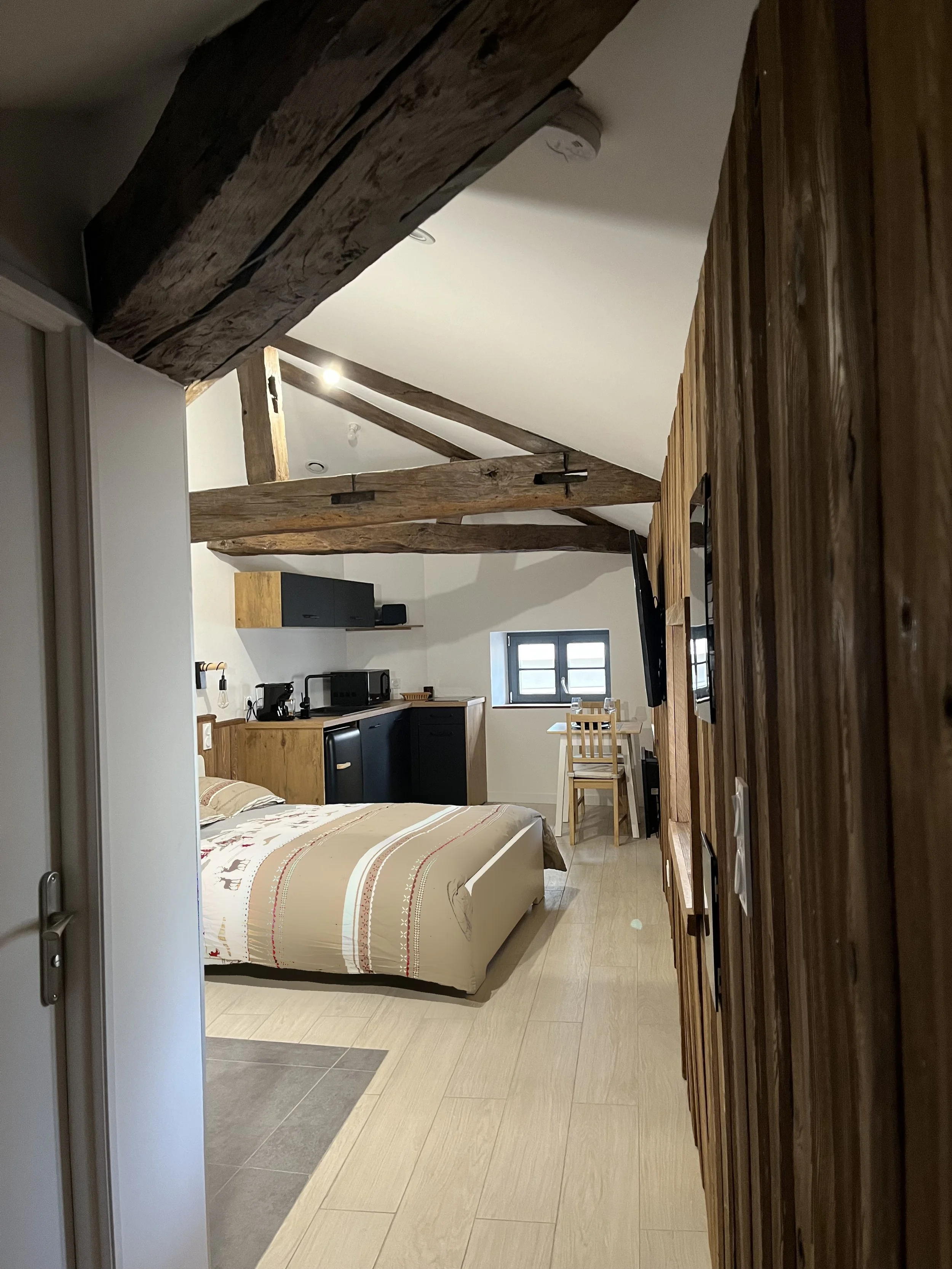 Chambre d'un appartement avec lit, cuisine, table, chaises, fenêtres, poutres en bois au plafond et mur en bois.