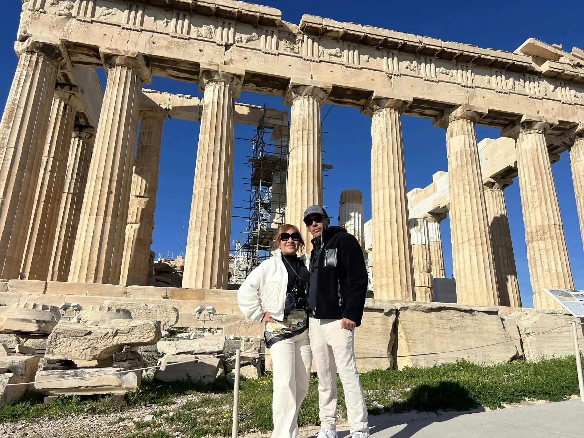Two tourists, a man and a woman, standing in front of the ancient Parthenon temple at the Acropolis in Athens, Greece. The Parthenon is partially under restoration with scaffolding visible, and the sky is clear and blue.