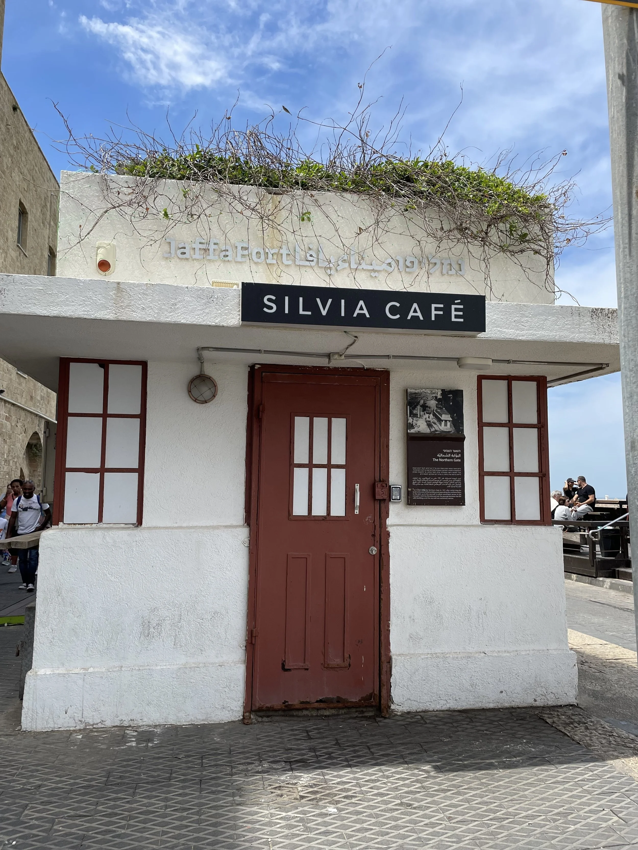 White building with a red door, black sign that says "Silvia Café," and Arabic text above the sign. The sky is partly cloudy. Several people are seen in the background near the right side.