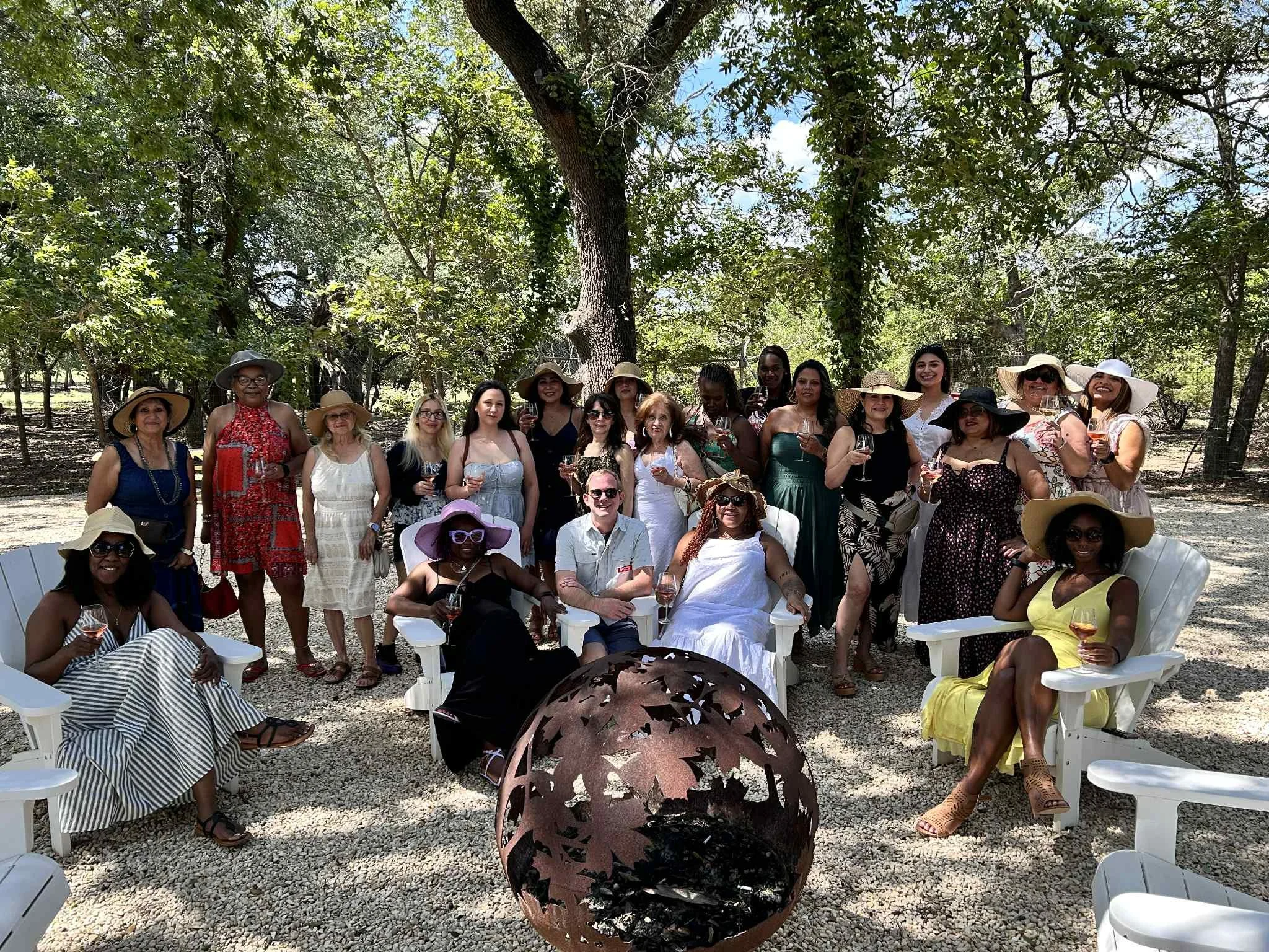 Group of women and men at outdoor gathering, seated on white plastic chairs, some holding wine glasses, in a wooded area under trees during daytime.