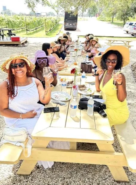 Group of women enjoying a summer outdoor meal at a long picnic table, wearing sun hats and sunglasses, with some holding drinks, in a sunny vineyard setting.
