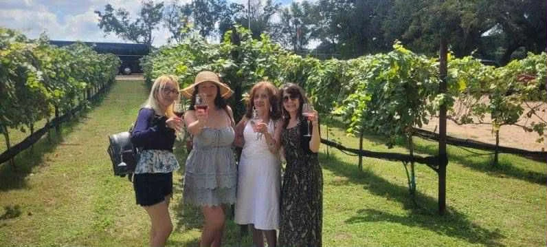 Four women standing together outdoors in a vineyard, smiling, with green vines and a parked car in the background.