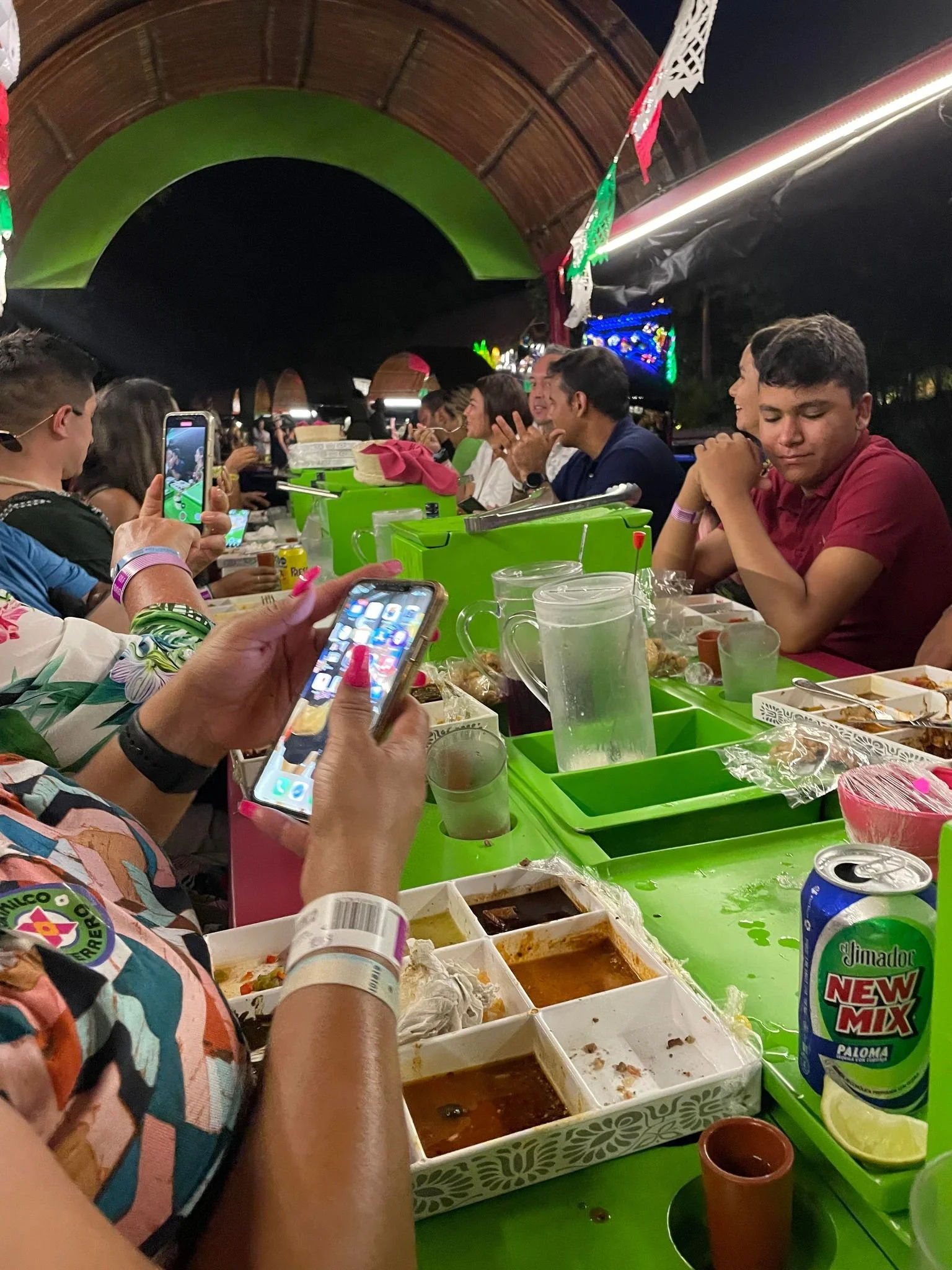 People sitting at a long table outdoors at night, eating food and taking photos, with a large green and brown canopy overhead and festive decorations hanging above.