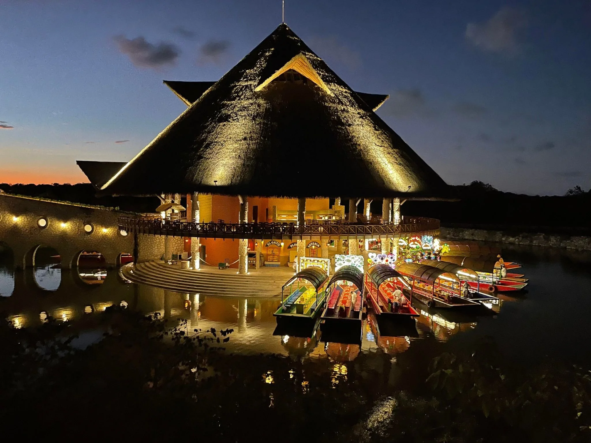 A large, thatched-roof building illuminated at night with decorative lighting, located near a water body with colorful pedal boats and an amusement park area in the foreground.