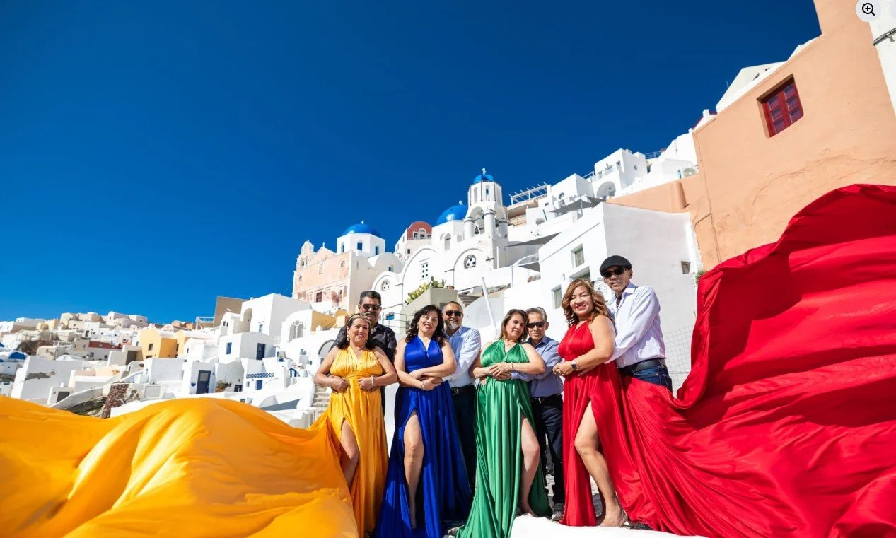 Group of people in colorful dresses standing in front of white buildings with blue domes in Santorini, Greece, under a clear blue sky.