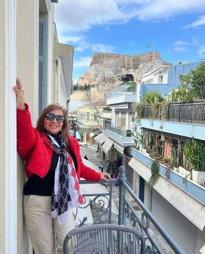 Woman in sunglasses and red jacket smiling on balcony with view of a street, pastel buildings, and a historic fortress on a hill in the background.