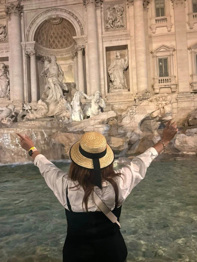 A woman wearing a straw hat and white shirt facing the Trevi Fountain in Rome, Italy, with her arms raised.