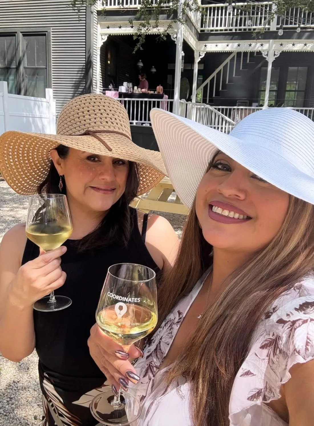 Two women wearing large sun hats are smiling and holding glasses of white wine. They are outdoors in a sunny setting with a house and patio visible in the background.