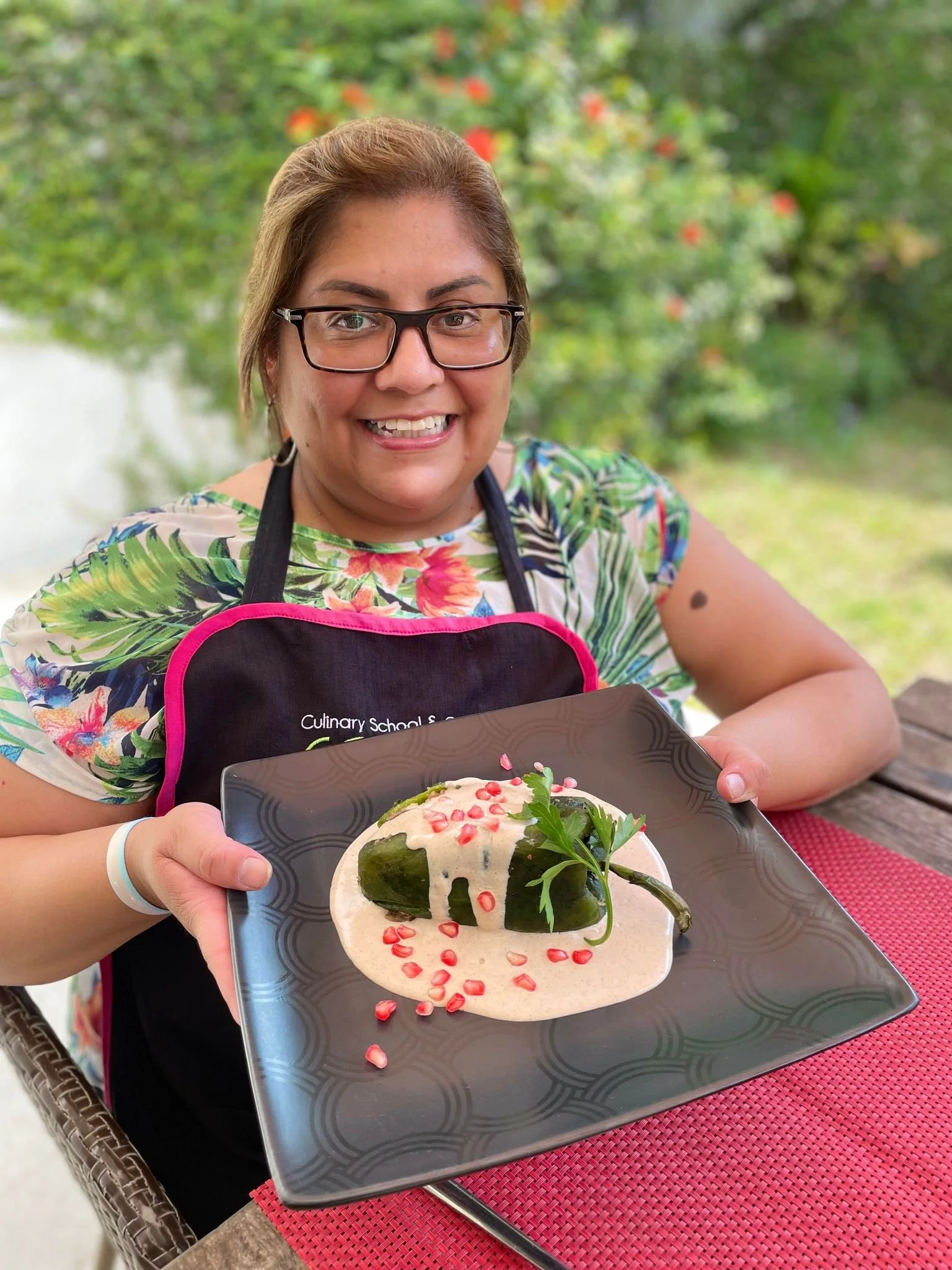 A woman with glasses holding a black serving plate with stuffed green chilies topped with pomegranate seeds and garnished with cilantro, sitting outdoors at a dining table.