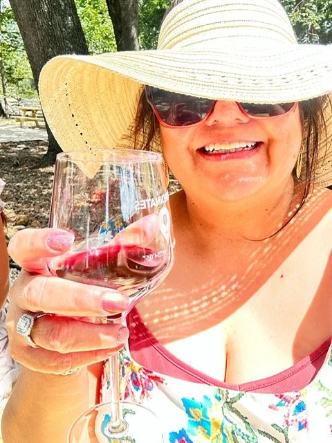 A woman outdoors wearing a large straw hat and sunglasses, smiling and holding a glass of red wine, with trees and sunlight in the background.