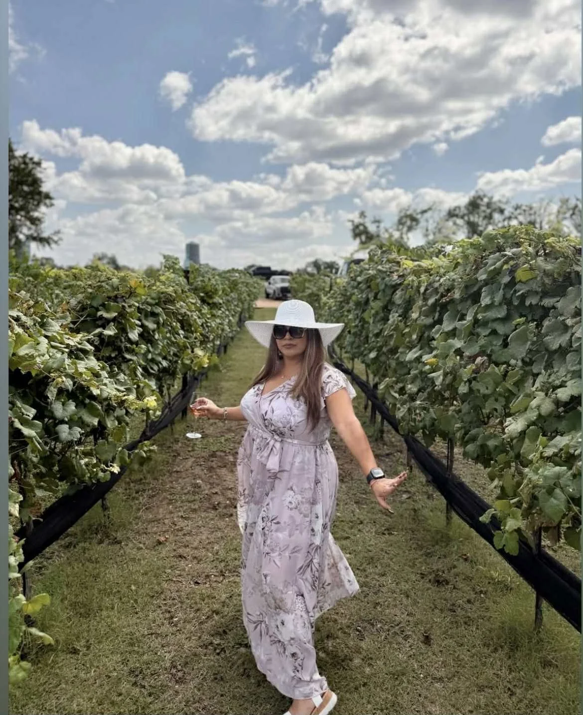 A woman standing in a vineyard aisle, wearing a floral dress, large white sun hat, sunglasses, and a smartwatch, with green vineyard rows on either side and a partly cloudy sky.