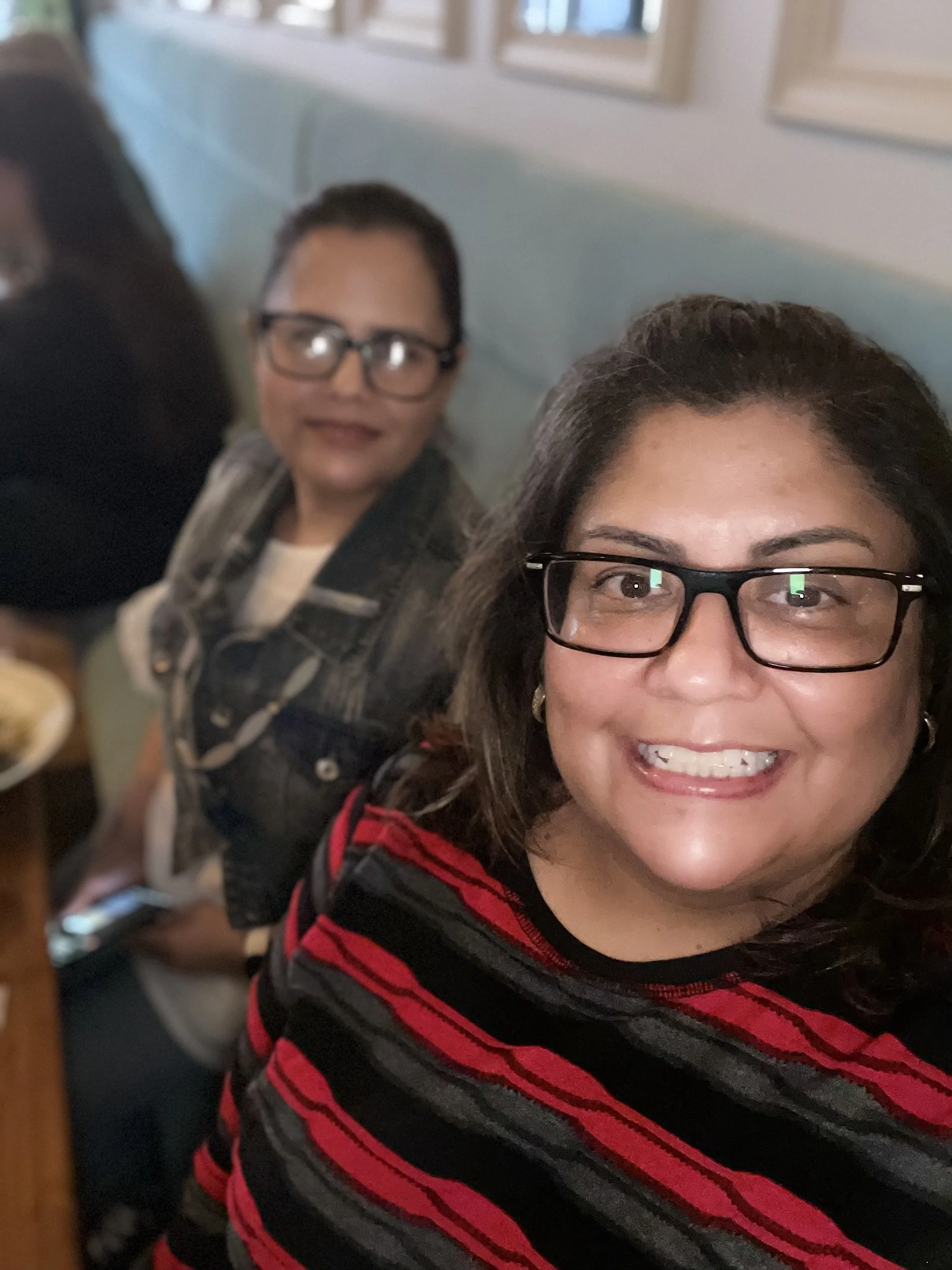 Close-up of a woman and a man in a restaurant, both wearing glasses and smiling, with the woman in the foreground and the man slightly in the background.