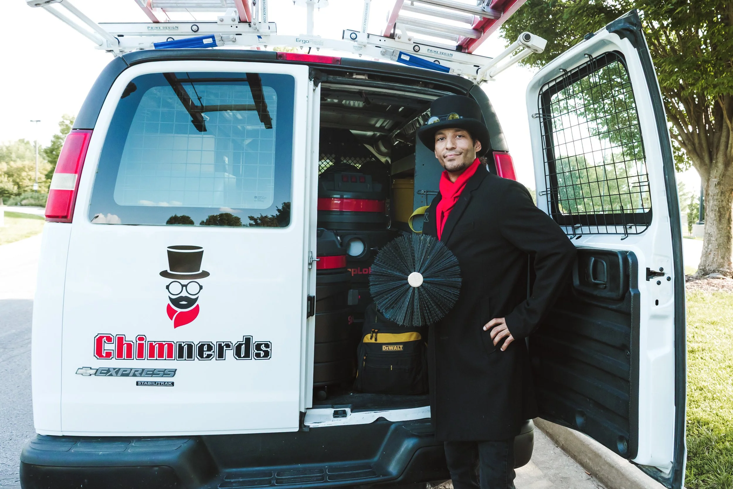 A man dressed as a chimney sweep in a black suit, black hat, and red scarf stands holding a chimney brush outside a white utility van with the logo and text "Chimnerds" on the side. 