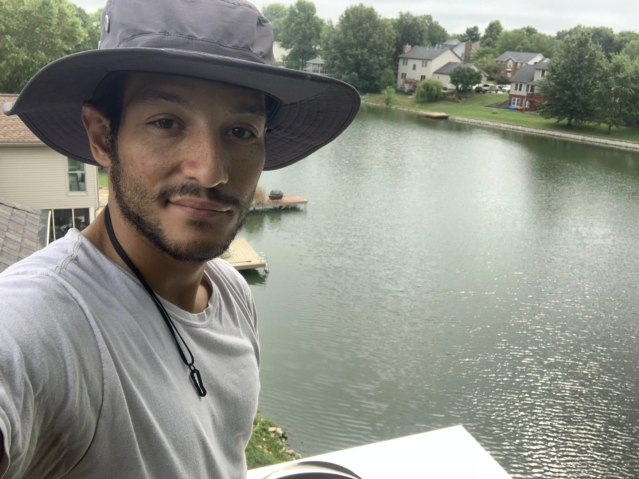 A young man wearing a wide-brimmed hat and gray shirt taking a selfie by a lake with houses and trees in the background.