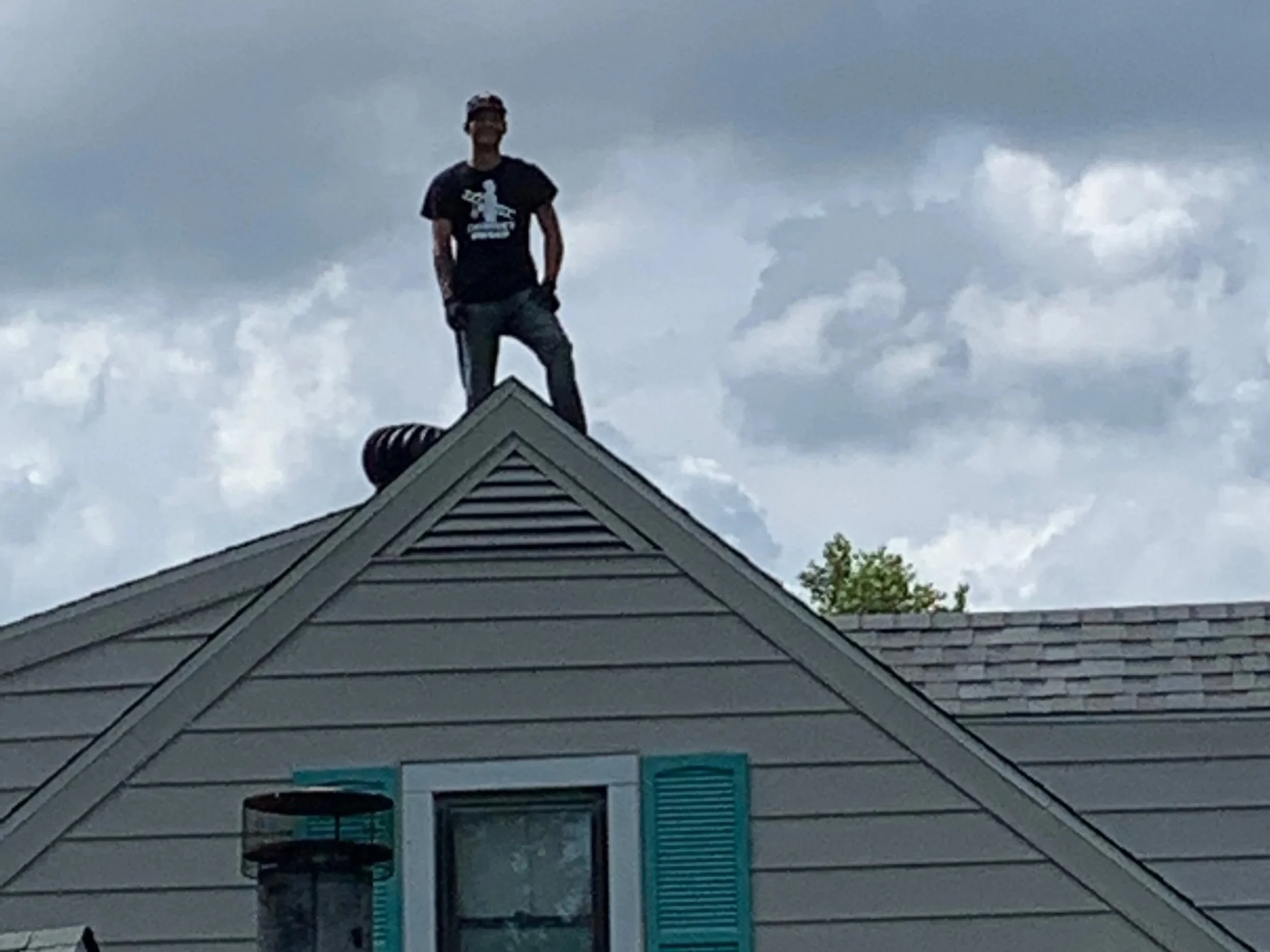 A man standing on the peak of a house roof against a cloudy sky.