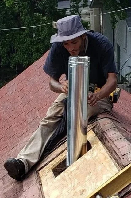 A man on a roof using a power tool next to a shiny metal vent pipe, wearing a gray hat and beige pants.