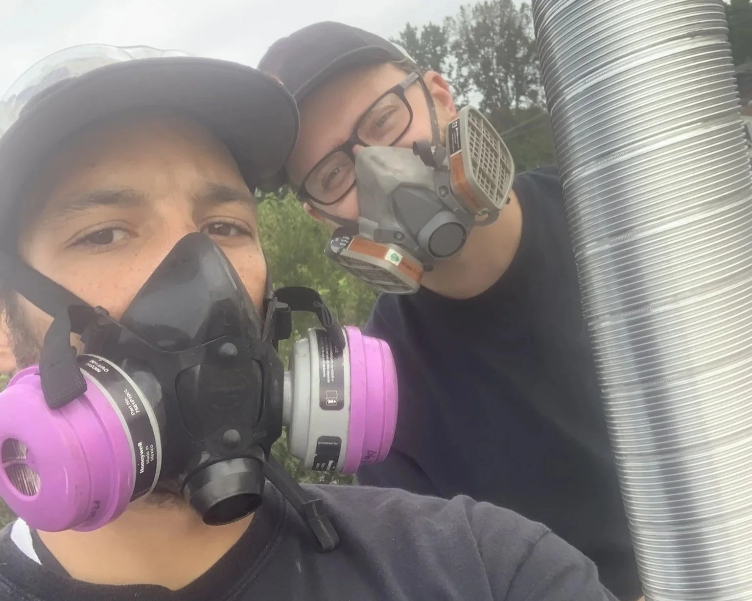 Two men wearing protective masks and safety glasses, one with a black mask and the other with a respirator, taking a selfie outdoors with trees in the background.
