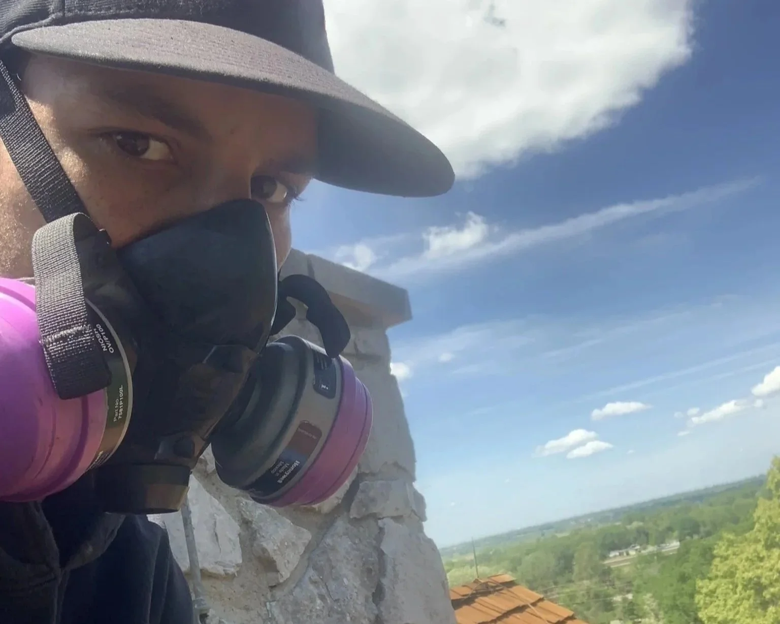 A person wearing a wide-brimmed hat, a face mask, and a pink respirator mask, appears to be a worker or climber on a rooftop with a landscape of trees and a partly cloudy sky in the background.