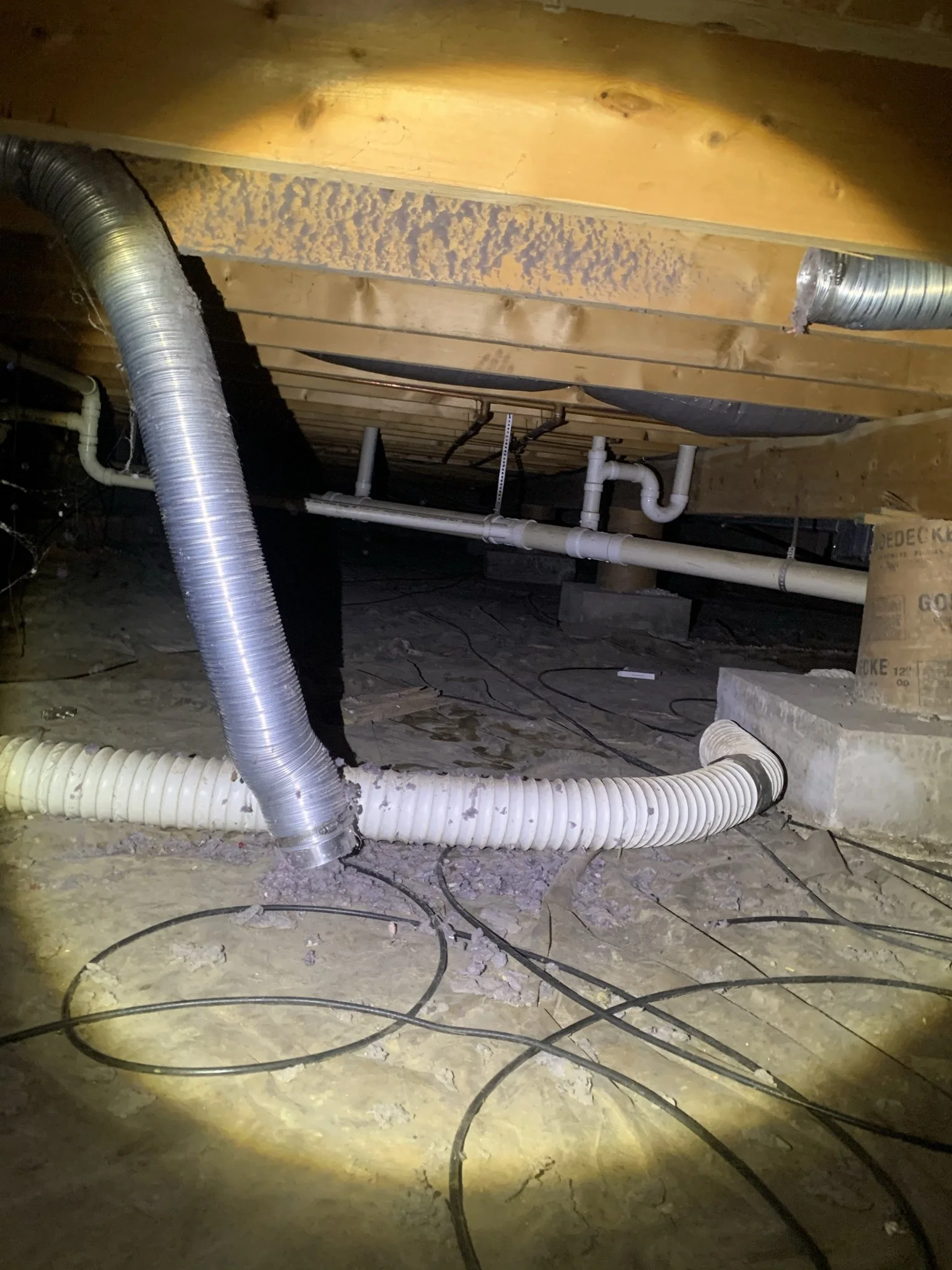 View of an unfinished basement or crawl space with exposed wooden beams, disconnected dryer vent ductwork, pipes, and electrical wires on a concrete floor.