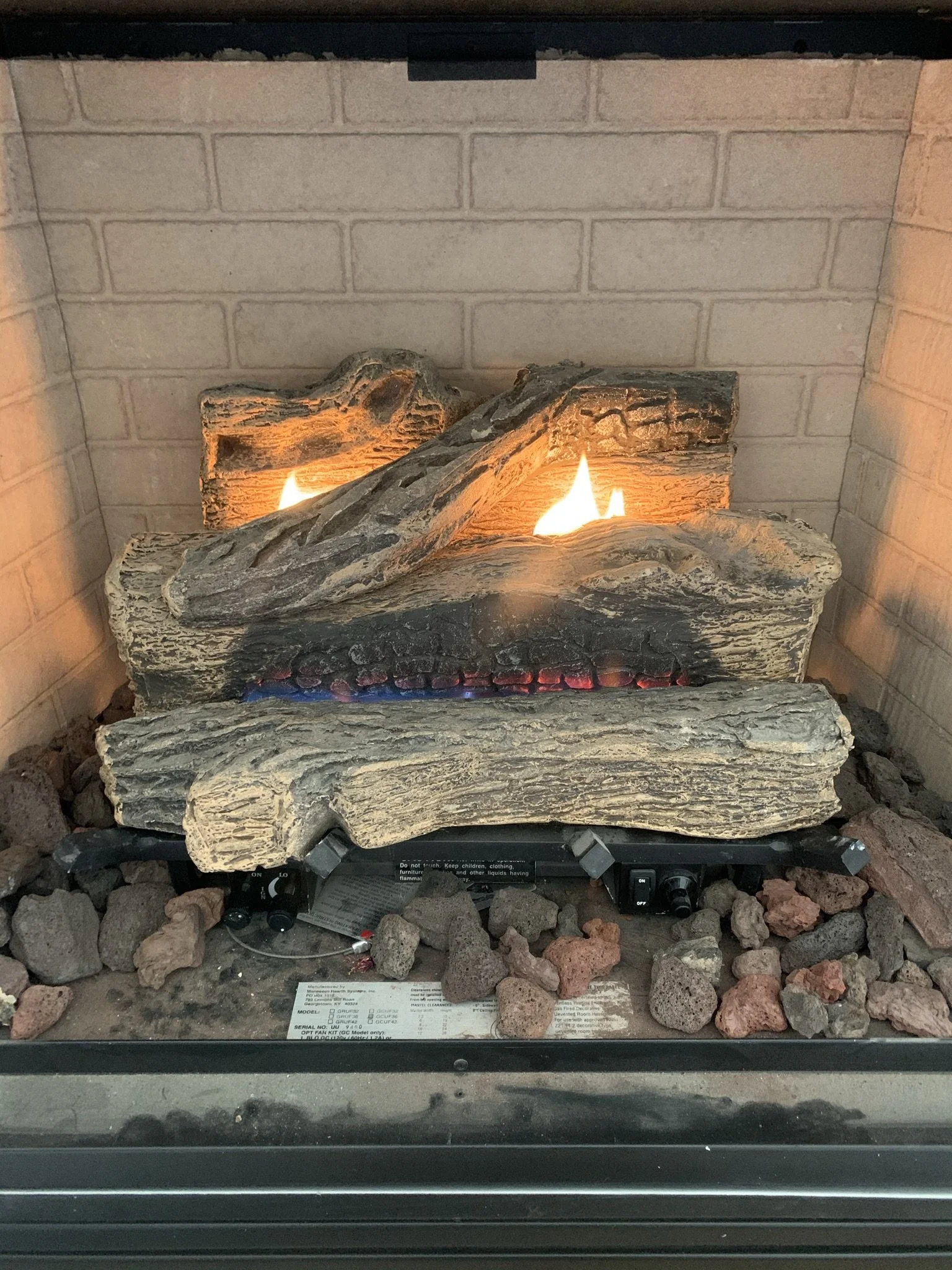 Electric fireplace with faux logs and flames, surrounded by black rocks and placed against a beige brick wall.