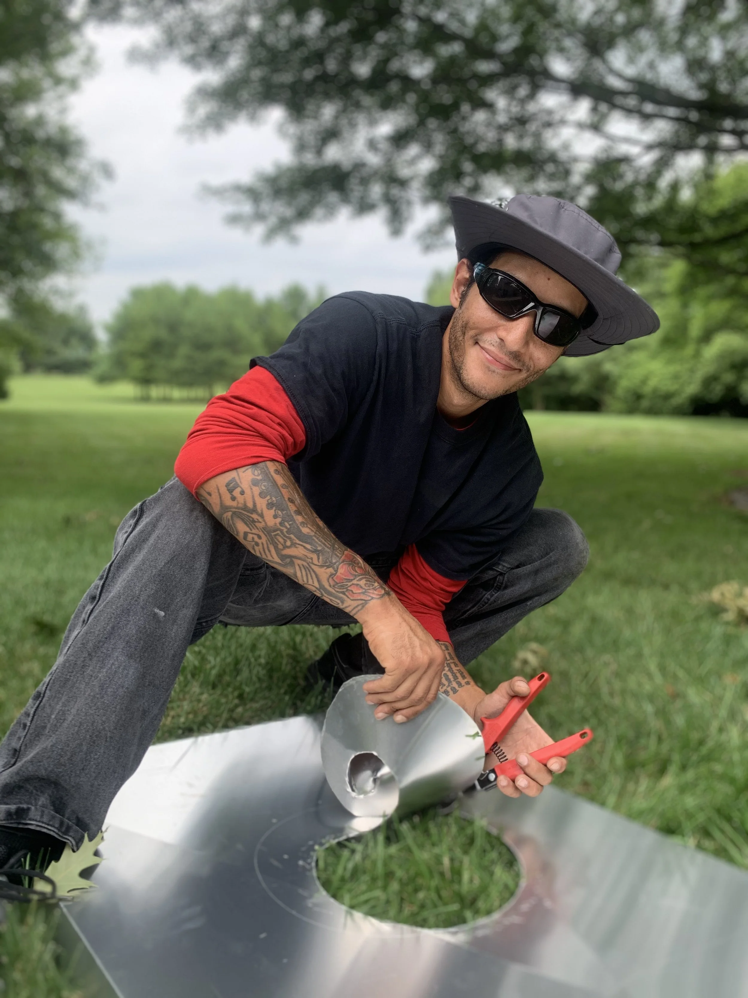 A man in sunglasses and a wide-brimmed hat working on a metal sculpture outdoors in a park-like setting.