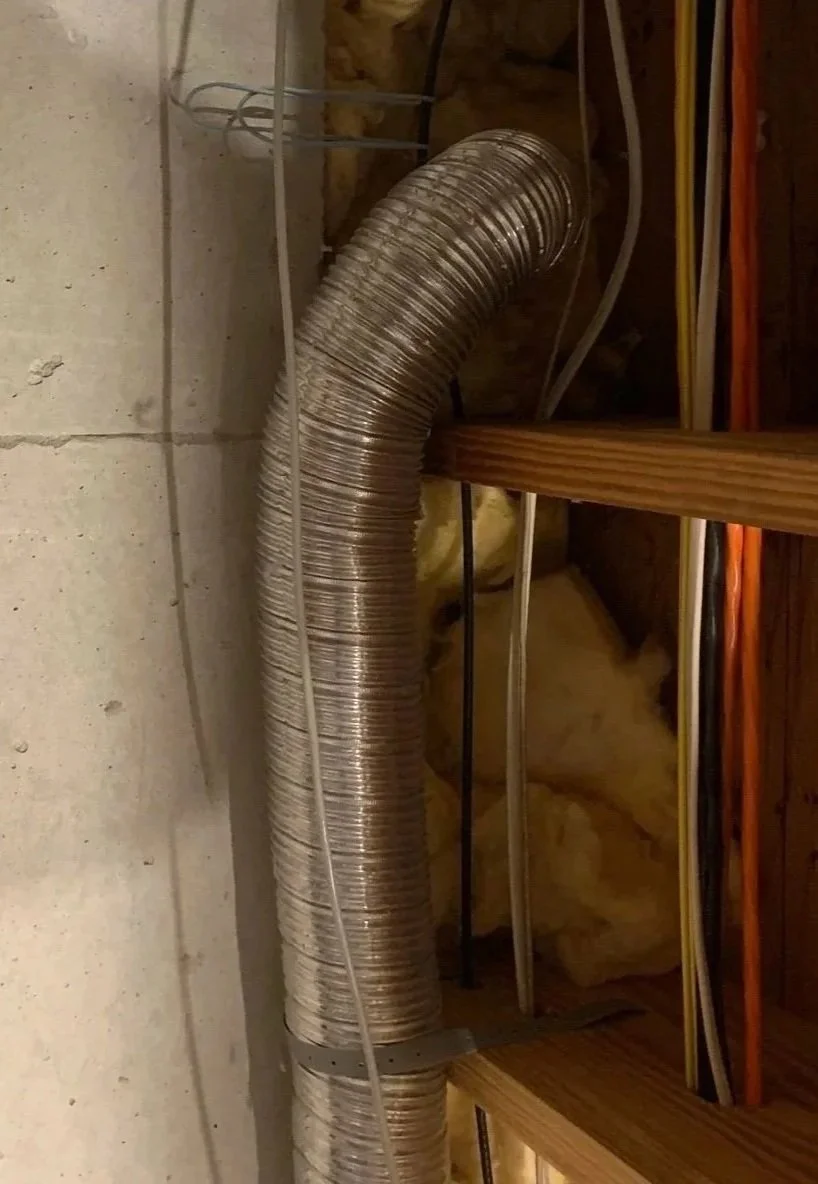 A flexible silver ventilation duct attached to a wooden beam in an unfinished basement or attic, surrounded by electrical wires and insulation.