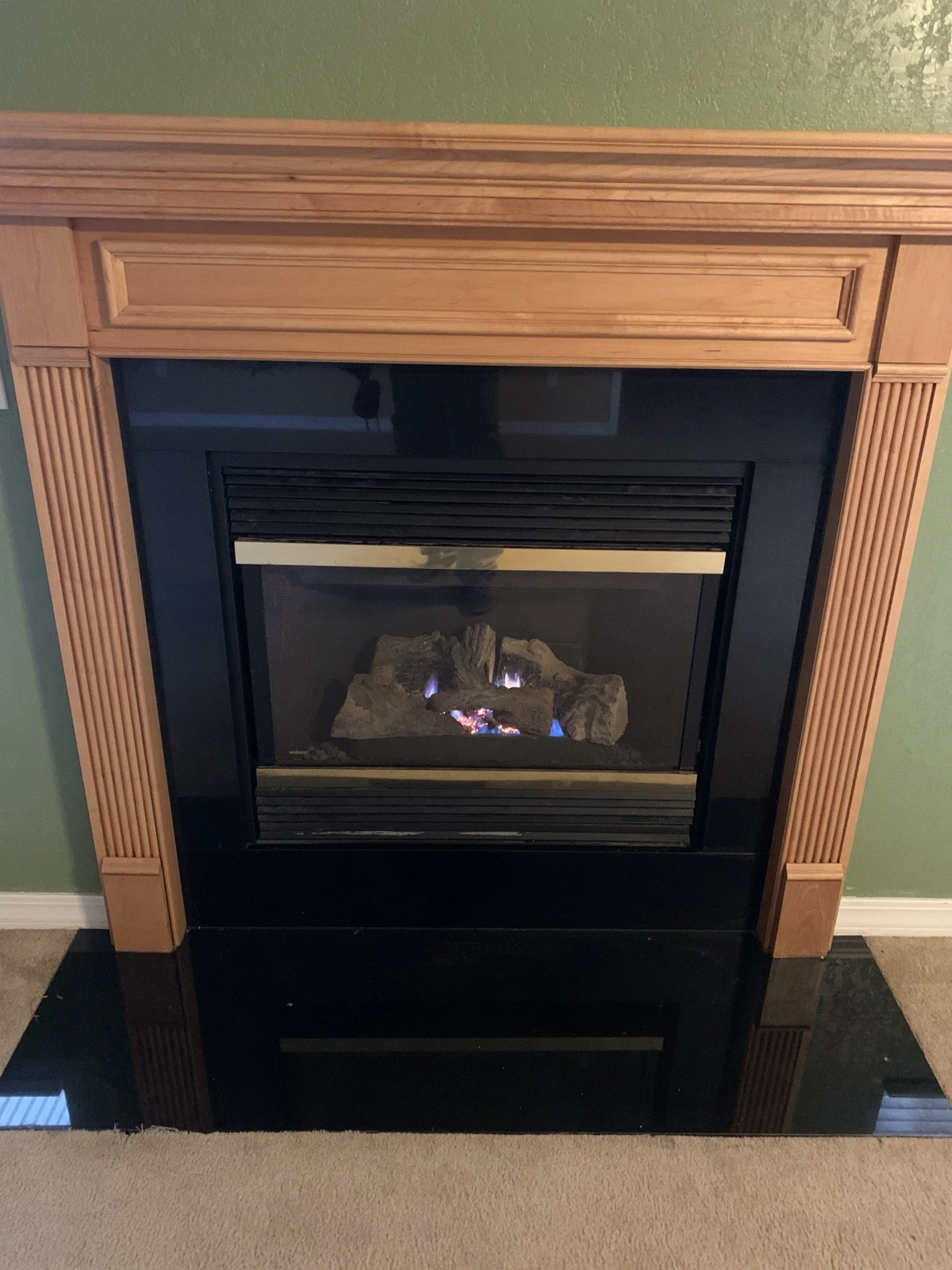 A fireplace with a wooden mantel, black glass surround, and gas logs, set against a green wall with a reflection on the black surface.