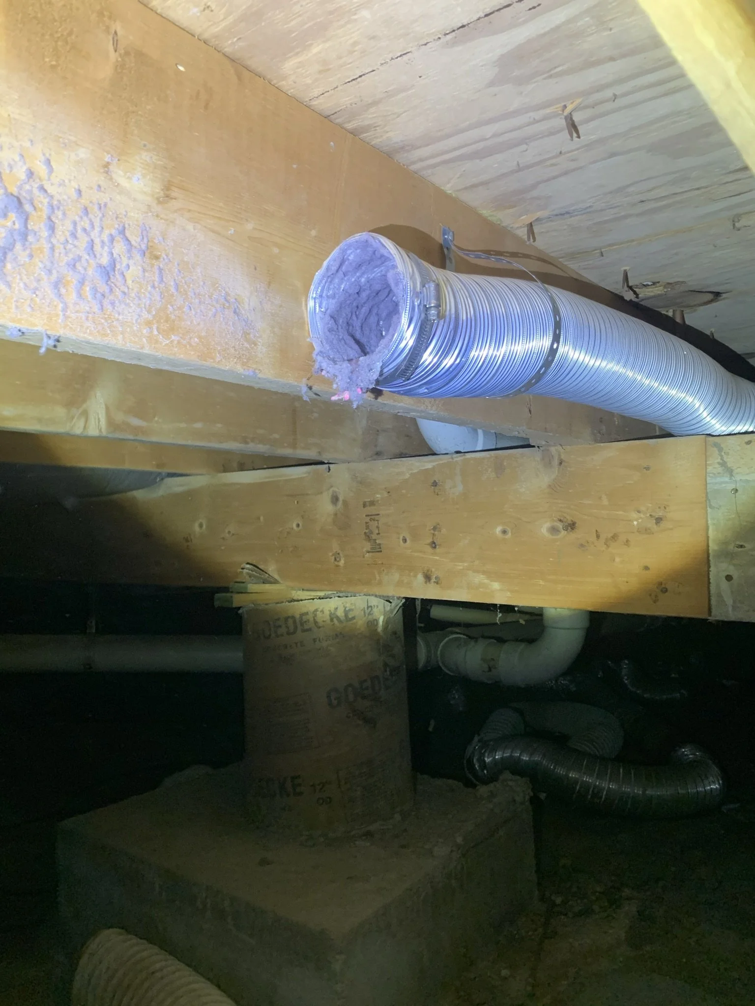 A construction or utility space with a large disconnected flexible metal duct, wiring, and wooden framing. The space appears to be part of a building under construction or renovation, with exposed pipes and electrical components.