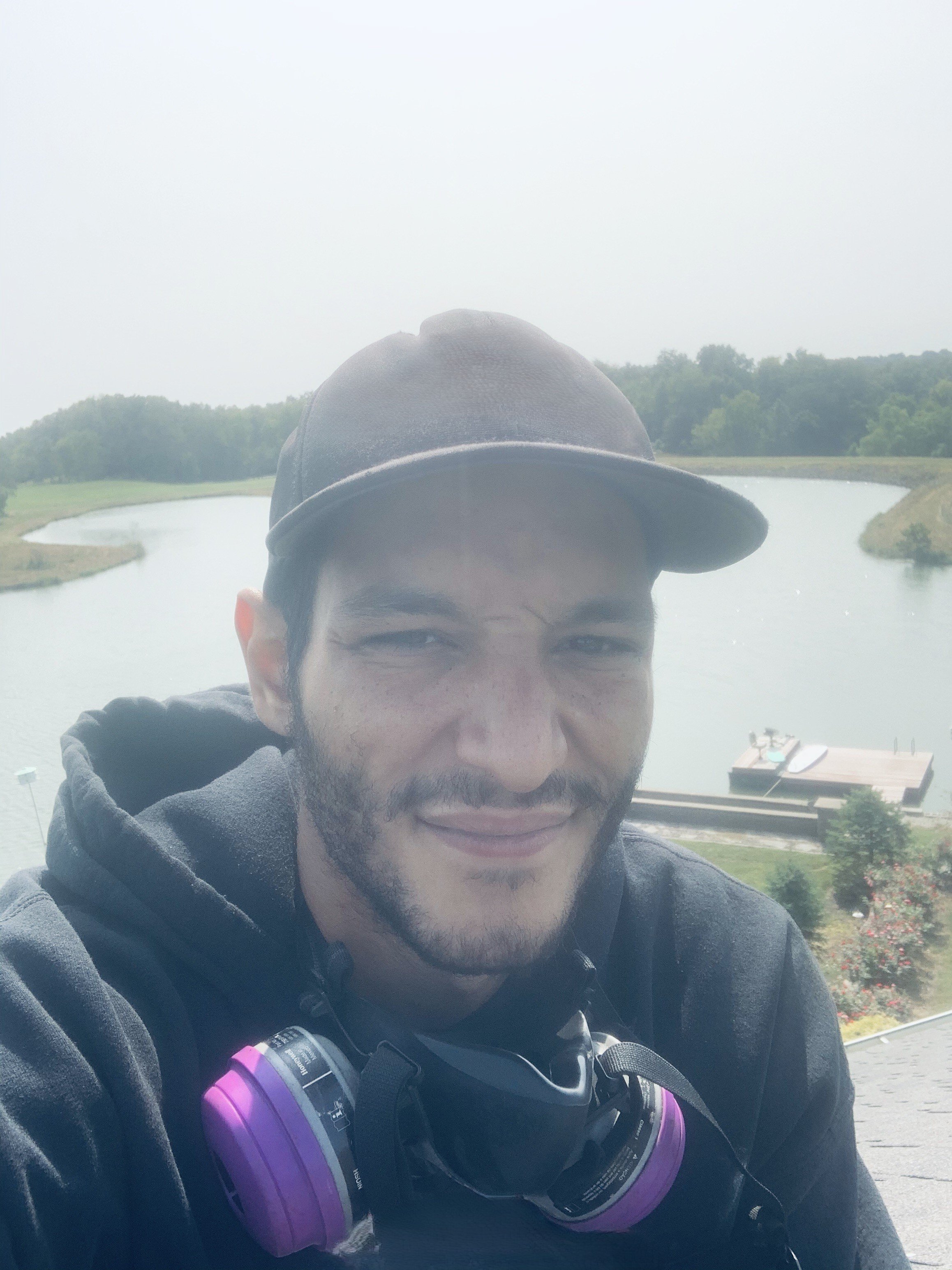 A man taking a selfie outdoors near a body of water, wearing a black cap, black hoodie, and purple and black headphones around his neck, with trees in the background.