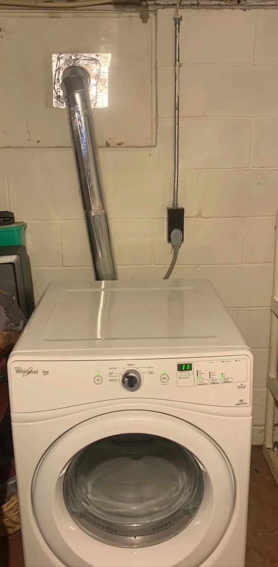 A front-loading dryer in a laundry room with rigid dryer vent connector pipe and brick wall background.