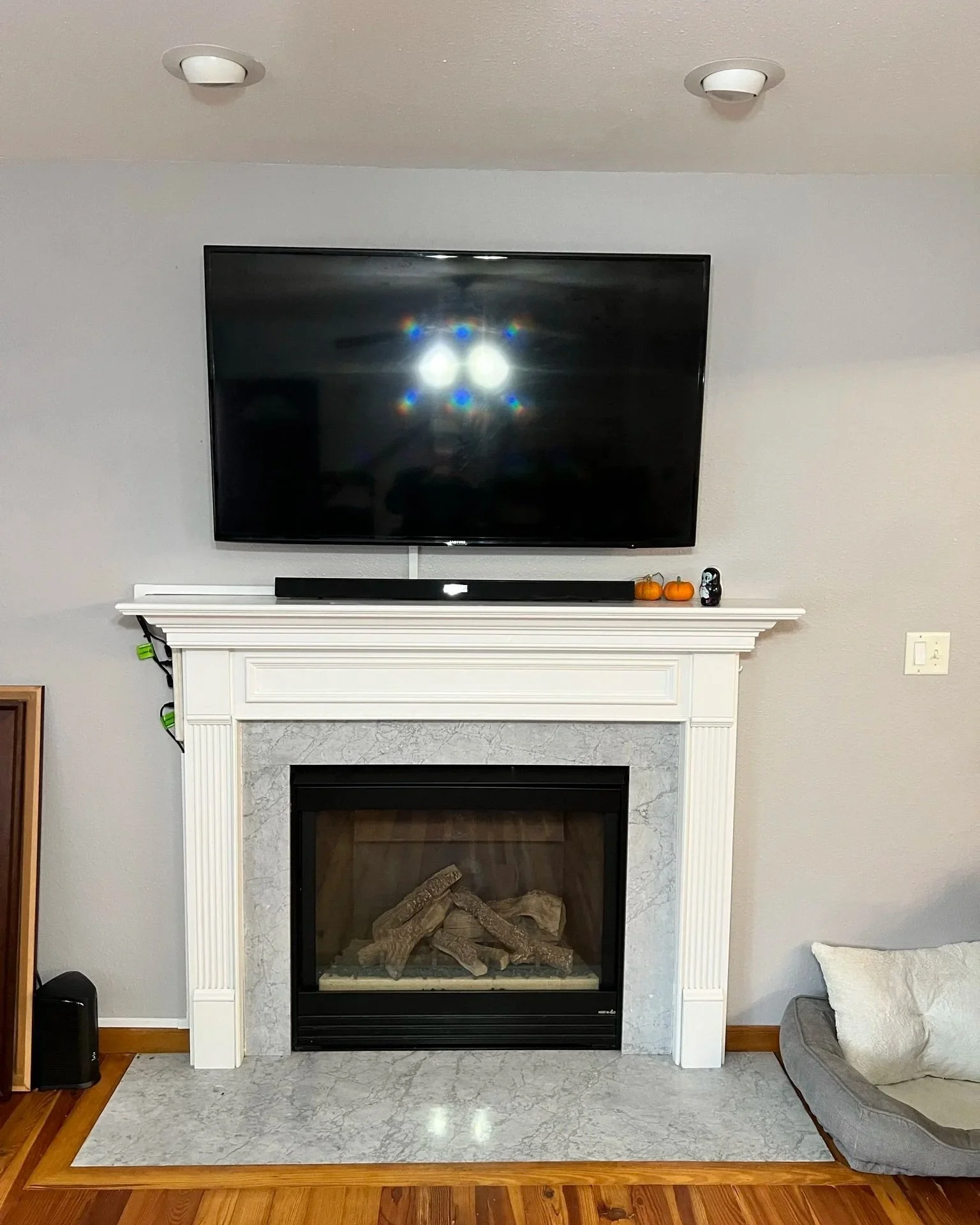 Living room fireplace with a mounted flat-screen TV above it, decorated with small pumpkins and a black skull on the mantel. A pet bed is on the right side of the fireplace.