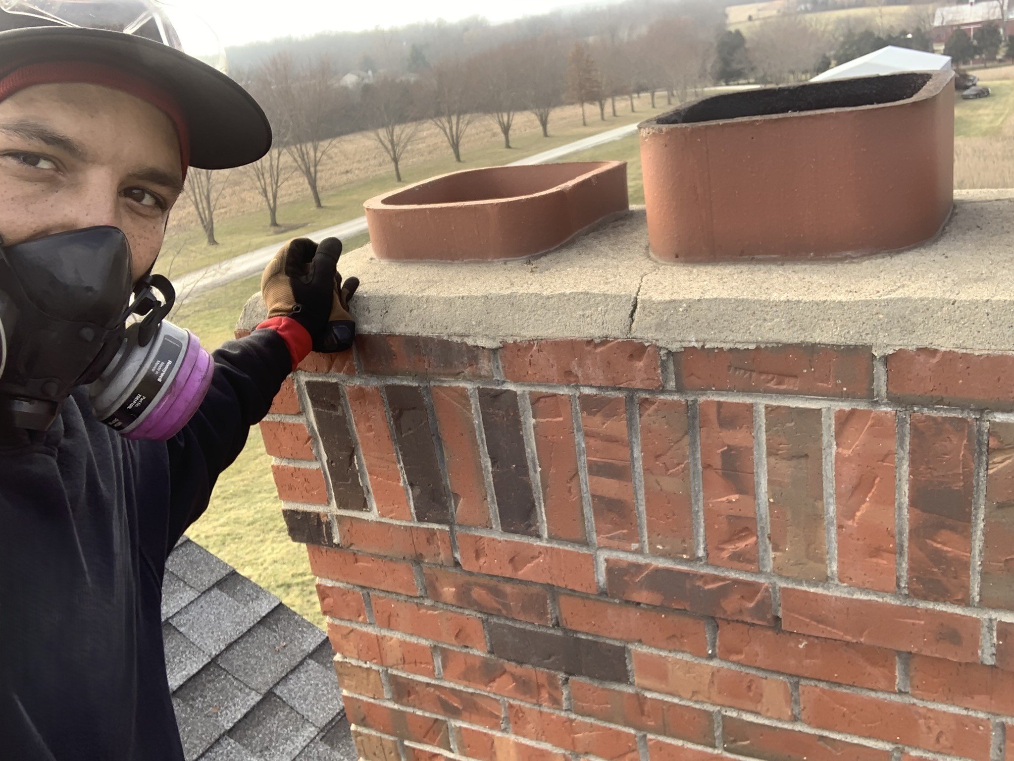A man wearing a cap, mask, and gloves is inspecting a brick chimney with two crossed clay chimney pots on top, during daytime with an overcast sky.