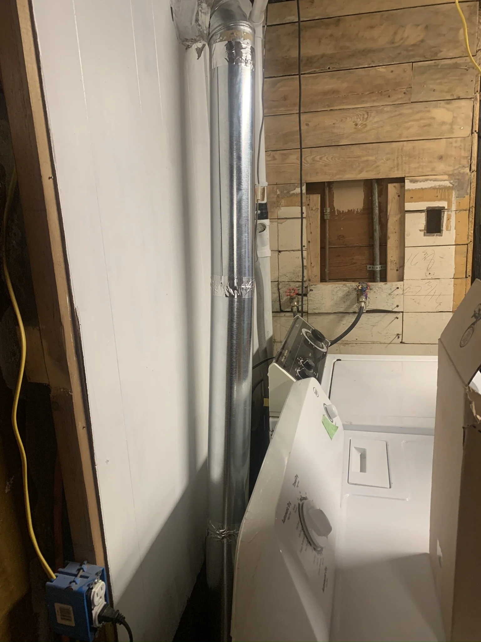 In a construction area, a shiny metallic rigid dryer vent pipe is installed on a white wall with nearby electrical outlets and wiring, and a dryer is situated in front of it.