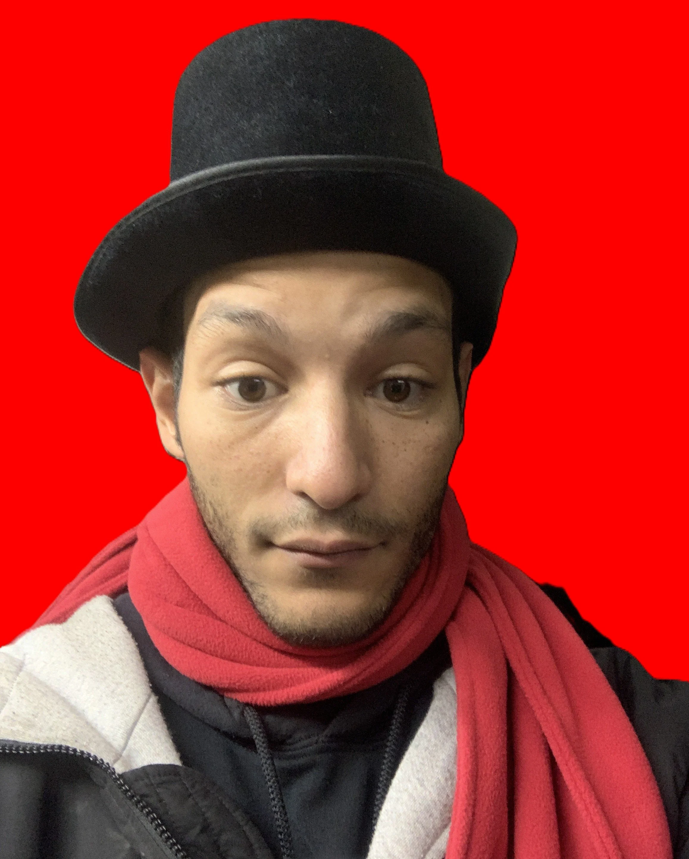 A young man wearing a black bowler hat and a red scarf, taking a selfie against a bright red background.